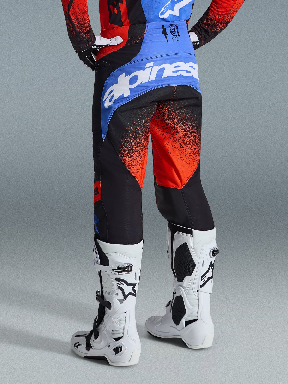Alpinestars 2026 Techstar Knif Pants, pantalon de motocross, orange, noir et bleu UCLA, vue arrière montrant l'empiècement bleu avec logo Alpinestars blanc, motif dégradé orange sur les jambes, porté avec un maillot assorti et des bottes blanches pour la course tout-terrain