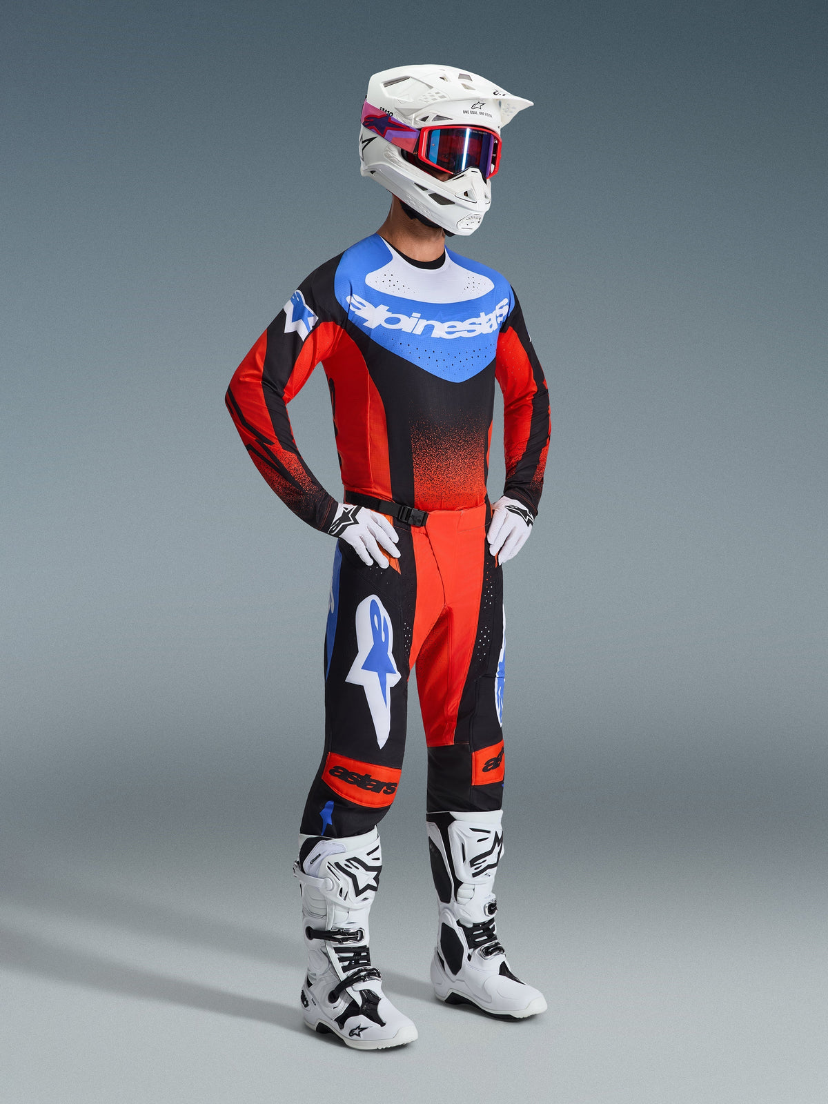 Pilote de motocross portant le pantalon Techstar Knif 2026 en orange, noir et bleu UCLA, avec un maillot de course assorti, un casque blanc avec des lunettes rouges et bleues et des bottes tout-terrain blanches, tenue professionnelle complète pour le motocross.