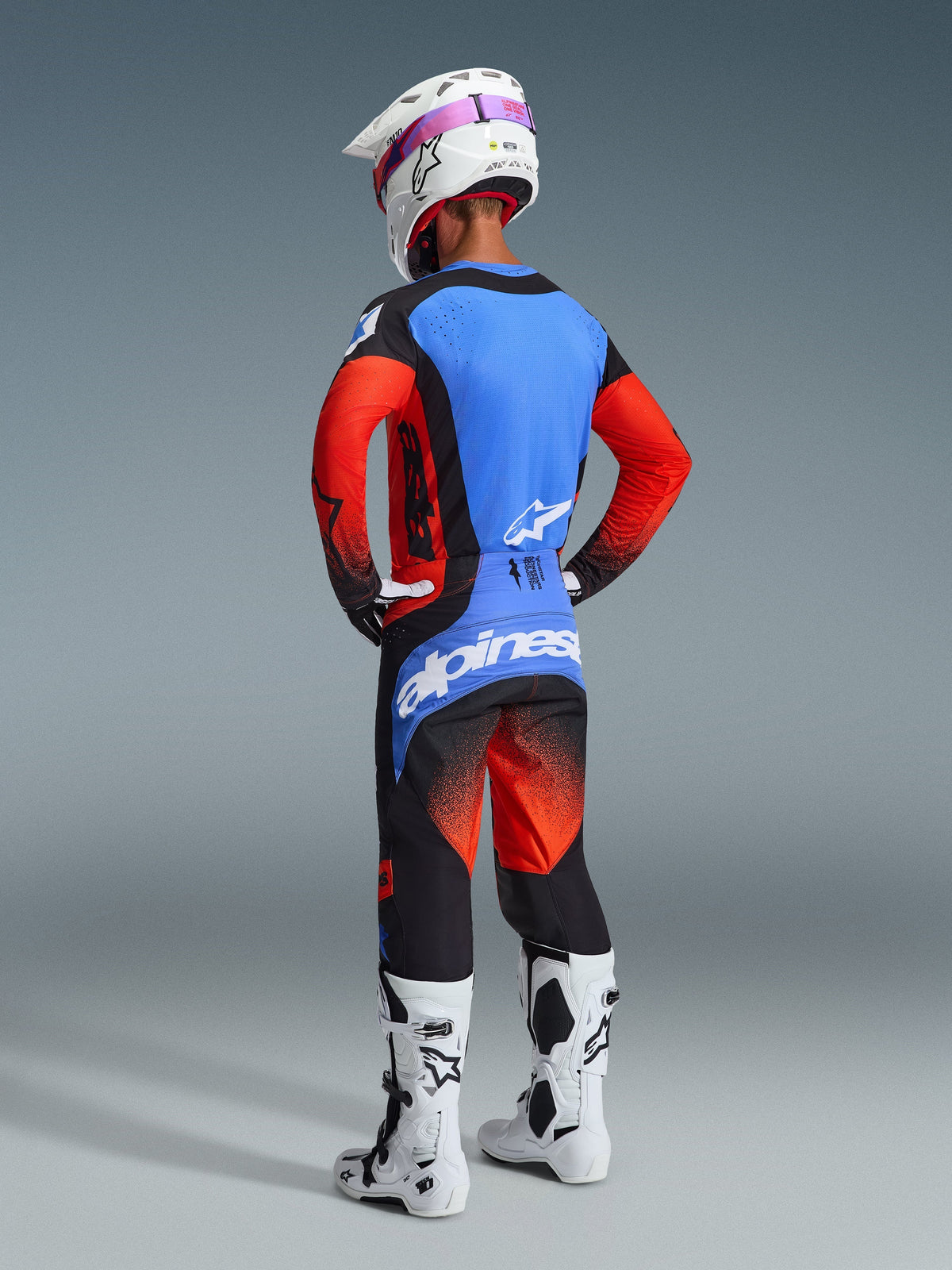 Pilote de motocross de dos portant une tenue de course complète, pantalon Techstar Knif 2026 en orange, noir et bleu UCLA, maillot bleu avec manches orange, casque blanc avec lunettes violettes, bottes blanches et noires, tenue coordonnée pour la performance tout-terrain