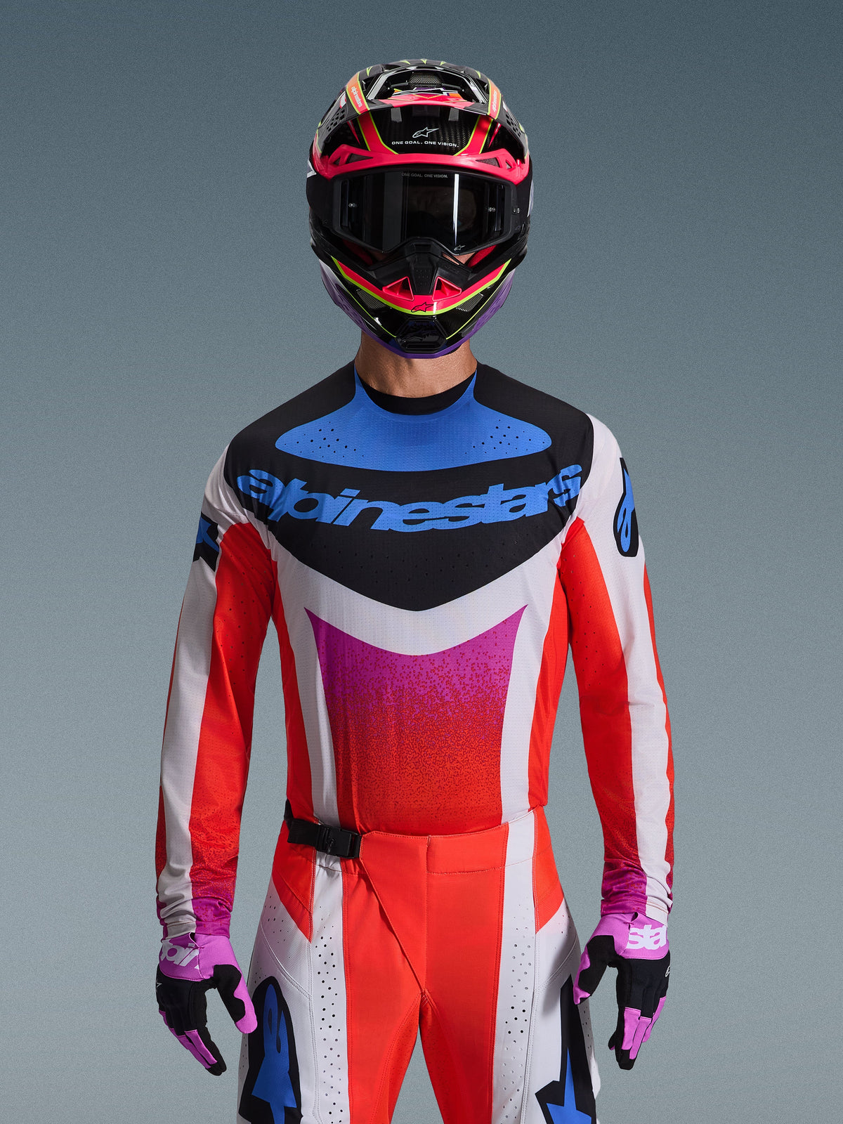 Motocross rider wearing the 2026 Techstar Knif Gear Set, white jersey with red sleeves and a blue and pink chest graphic, matching red and white pants, vibrant pink and yellow helmet with dark goggles, and purple and black gloves, professional gear for off-road racing.