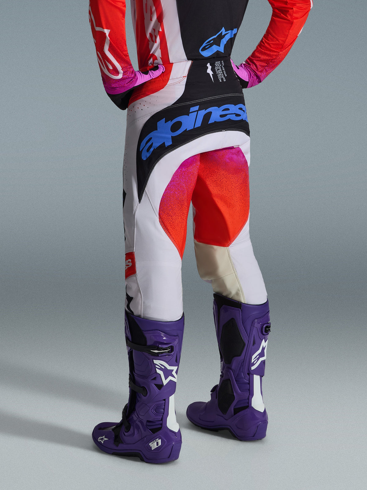 Alpinestars 2026 Techstar Knif Pants and Gear Set, Rear view of a motocross rider wearing white pants with black panels, pink and red gradient accents, and a large blue Alpinestars logo on the seat, paired with purple Tech 10 boots and a matching red and black jersey.
