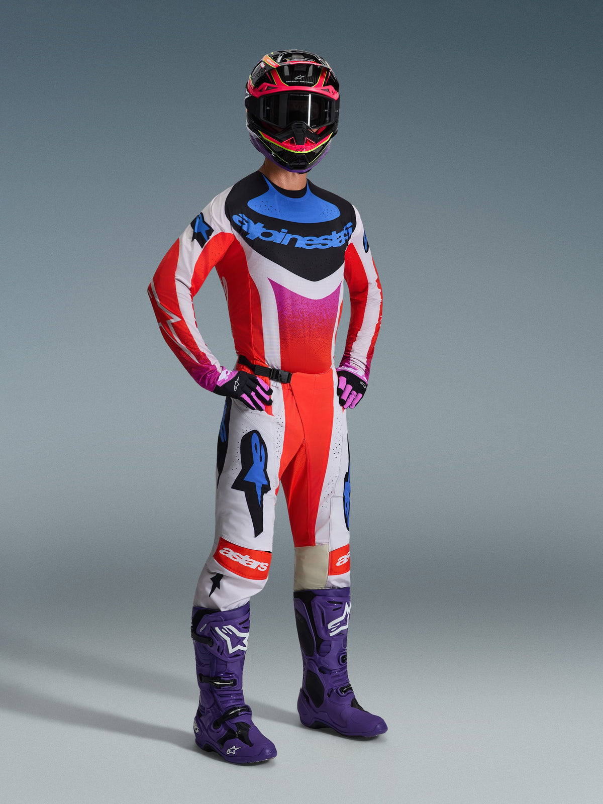 Alpinestars 2026 Techstar Knif Gear Set, Motocross rider wearing full racing gear, red jersey with white and blue accents, white pants with red and blue star logos, purple motocross boots, black and pink helmet, professional off-road racing kit