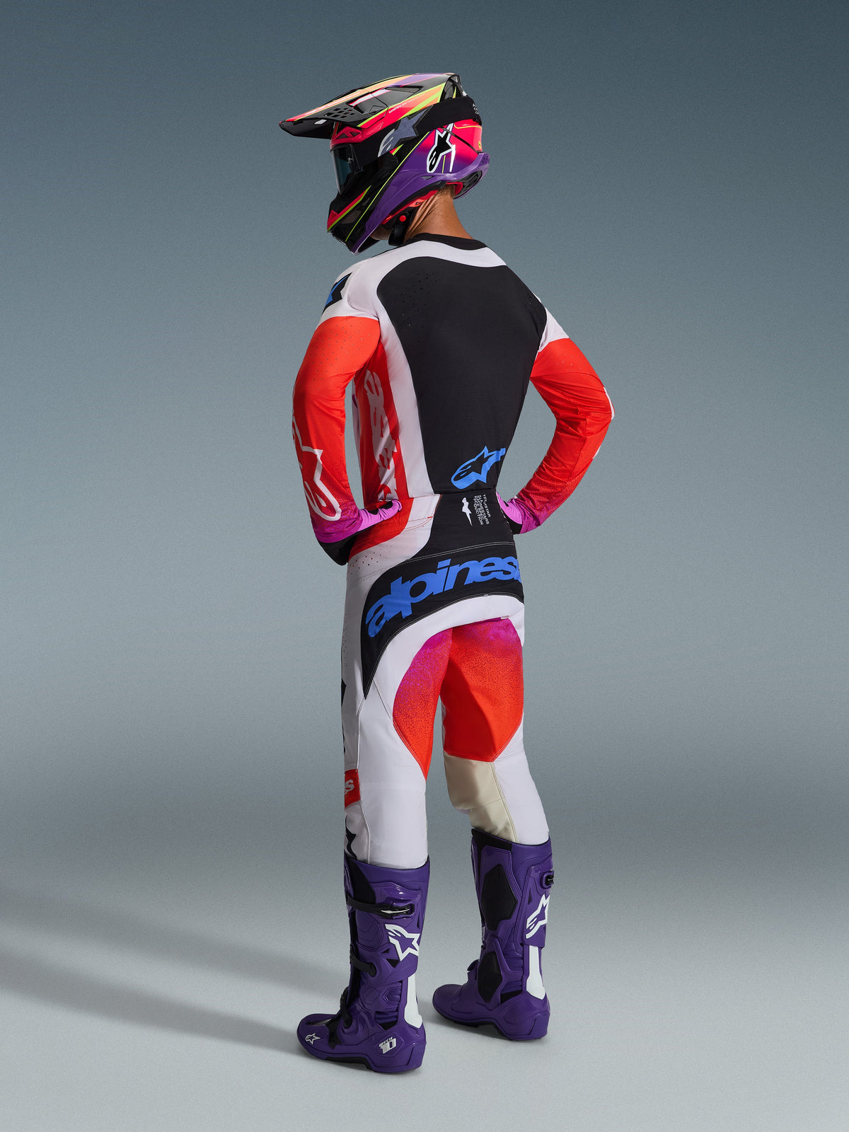 Motocross rider from the rear wearing the Alpinestars 2026 Techstar Knif Gear Set, white jersey with red sleeves and black panel, white pants with red and purple accents and blue Alpinestars branding, purple racing boots and matching helmet, professional off-road racing kit