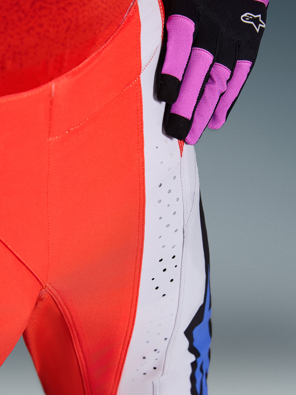 Alpinestars 2026 Techstar Knif Pants, close-up of hip and side panel in Orange Fluo and Light Gray, featuring laser-cut perforations for airflow, worn with a black and purple motocross glove, high-performance racing gear