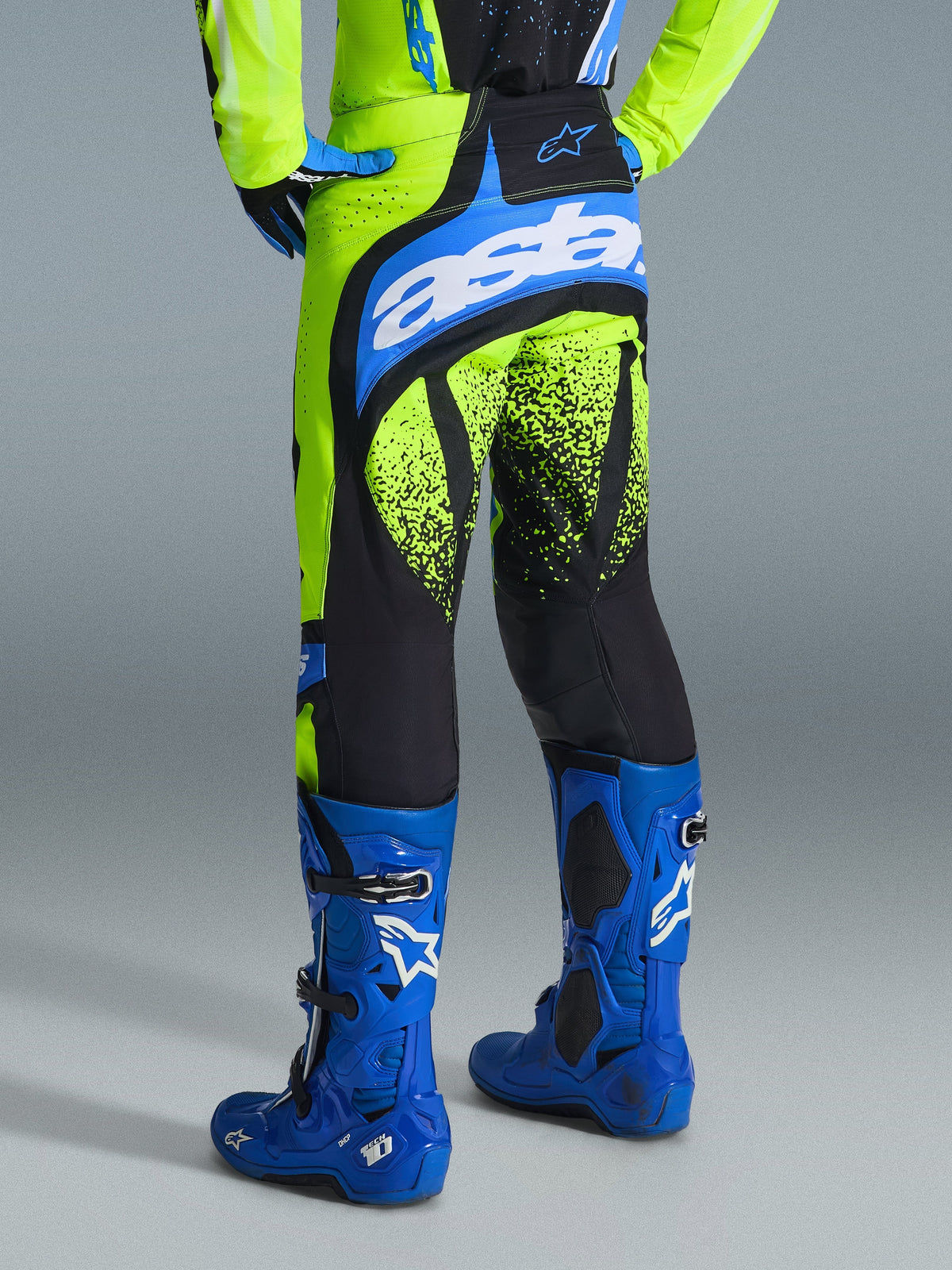 Alpinestars Pantalon Techstar Nomur 2026, pantalon de motocross, Jaune Fluo et Bleu UCLA, vue arrière du pilote portant des bottes bleues, avec logo « astars » sur l'assise et motifs d'éclaboussures noires sur les jambes