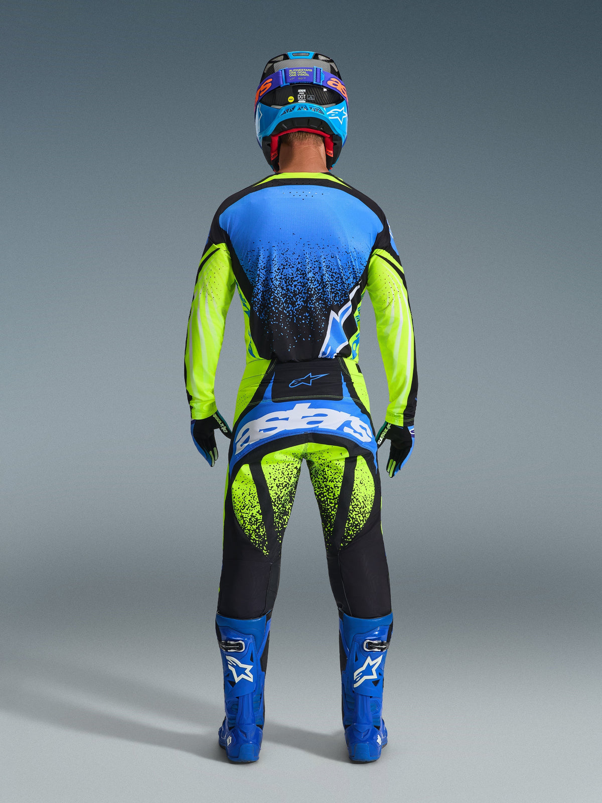 Pilote de motocross vu de dos portant le pantalon Alpinestars Techstar Nomur 2026, jaune fluo et bleu UCLA avec accents bleu marine foncé, maillot assorti et bottes bleues, avec logo Astars imposant sur l'assise et motif graphique moucheté.