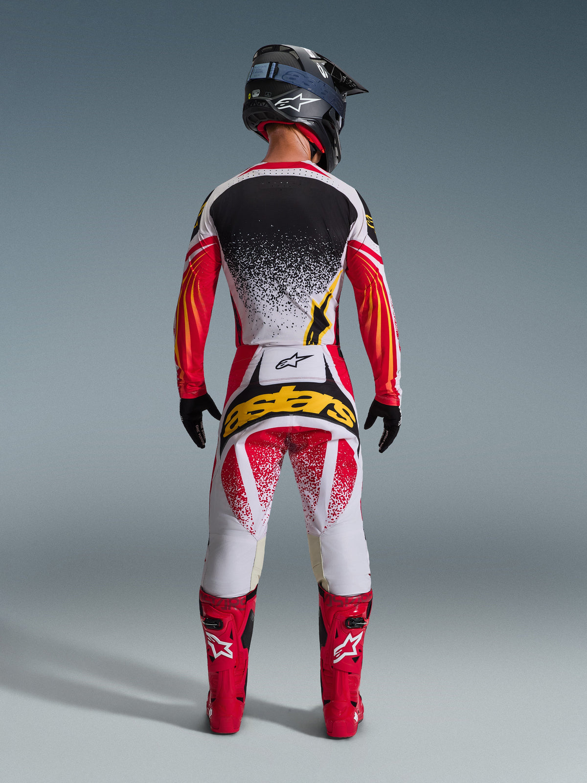 Ensemble d'équipement Alpinestars Techstar Nomur 2026, tenue de motocross complète vue de dos, maillot blanc avec effet d'éclaboussures noires et manches aux graphismes rouge-orange, pantalon blanc et rouge avec logo 'astars' jaune, bottes de course rouges, casque noir texture carbone avec lunettes bleues, équipement tout-terrain haute performance.