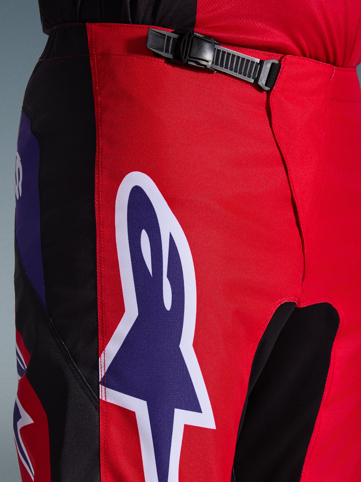 Alpinestars 2026 Fluid Grid Pants, Motocross Pants, Red Black and Purple, close-up of the waist and hip area featuring a black ratchet closure system and a large purple Alpinestars star logo with white outline, durable textured fabric for off-road racing.