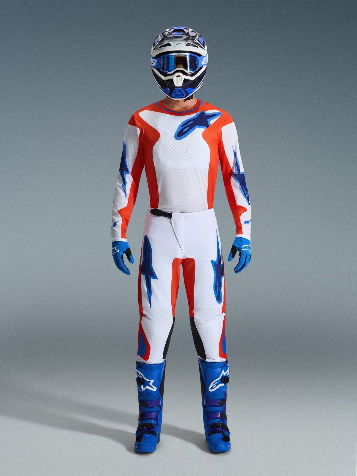 Motocross rider wearing complete racing gear, Alpinestars 2026 Fluid Grid Pants in Orange and UCLA Blue, white jersey with orange sleeves and blue logos, matching blue helmet and goggles, blue boots and blue gloves, coordinated outfit for off-road racing