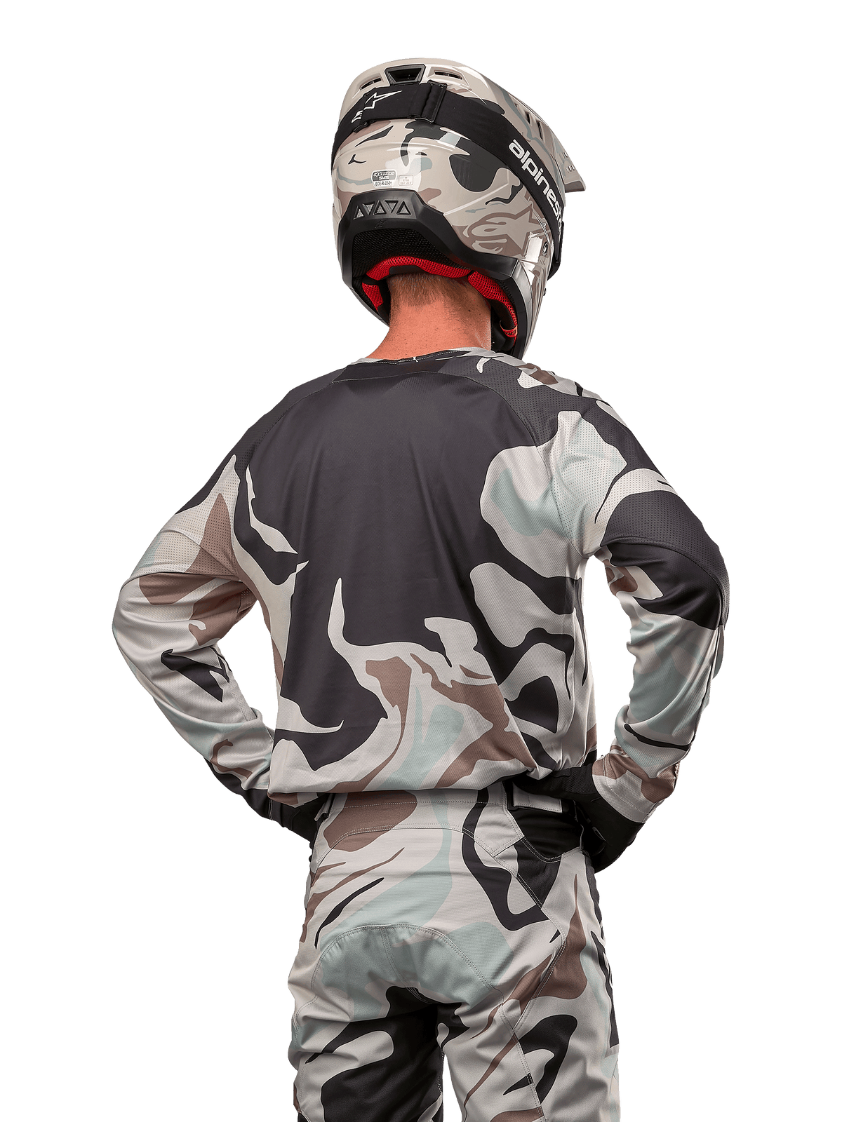 Pilote de motocross vu de dos portant le maillot Alpinestars Racer Tactical 2024 en Iron Camo Dust Gray, pantalon et casque camouflage assortis, mains sur les hanches, équipement de course tout-terrain coordonné.