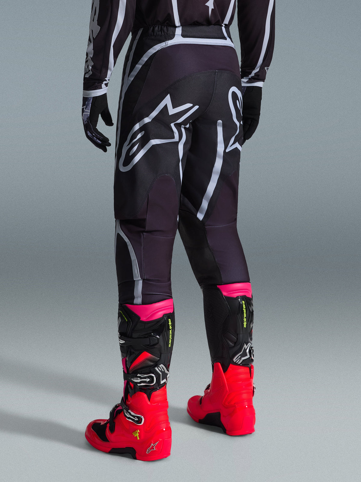 Alpinestars Fluid Apex Pants 2026, pantalon de motocross, noir et gris, vue arrière présentant de grands logos Alpinestars blancs sur l'assise, porté avec des bottes rouge vif et rose, construction durable en poly-tissu 600D pour des performances tout-terrain