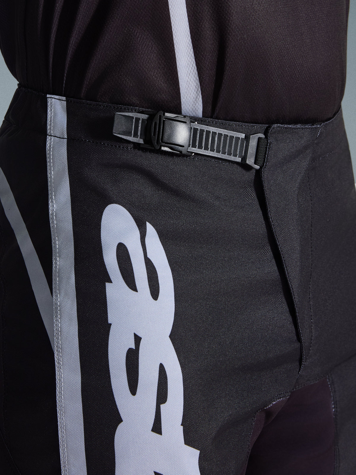 Pantalon Alpinestars Fluid Apex 2026, pantalon de motocross, noir et gris, gros plan sur le système de fermeture à cliquet intégré pour un ajustement sécurisé, avec graphismes de logo blancs sur un poly-tissu 600D durable