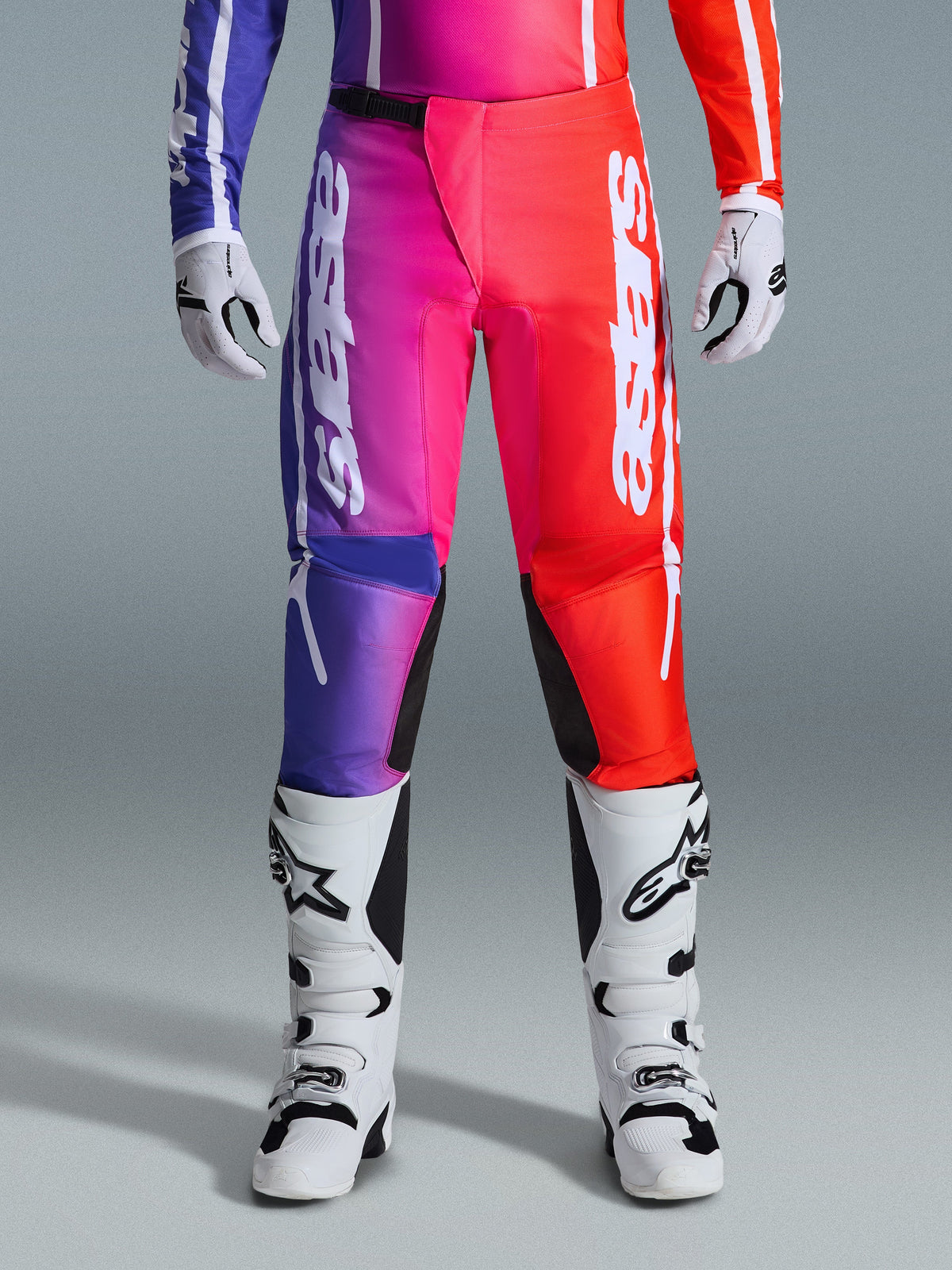 Alpinestars 2026 Fluid Apex Pants, pantalon de motocross, design dégradé multicolore violet, rose et rouge, grand marquage Alpinestars blanc sur les jambes, associé à des bottes et des gants de motocross blancs et noirs, équipement de course tout-terrain professionnel
