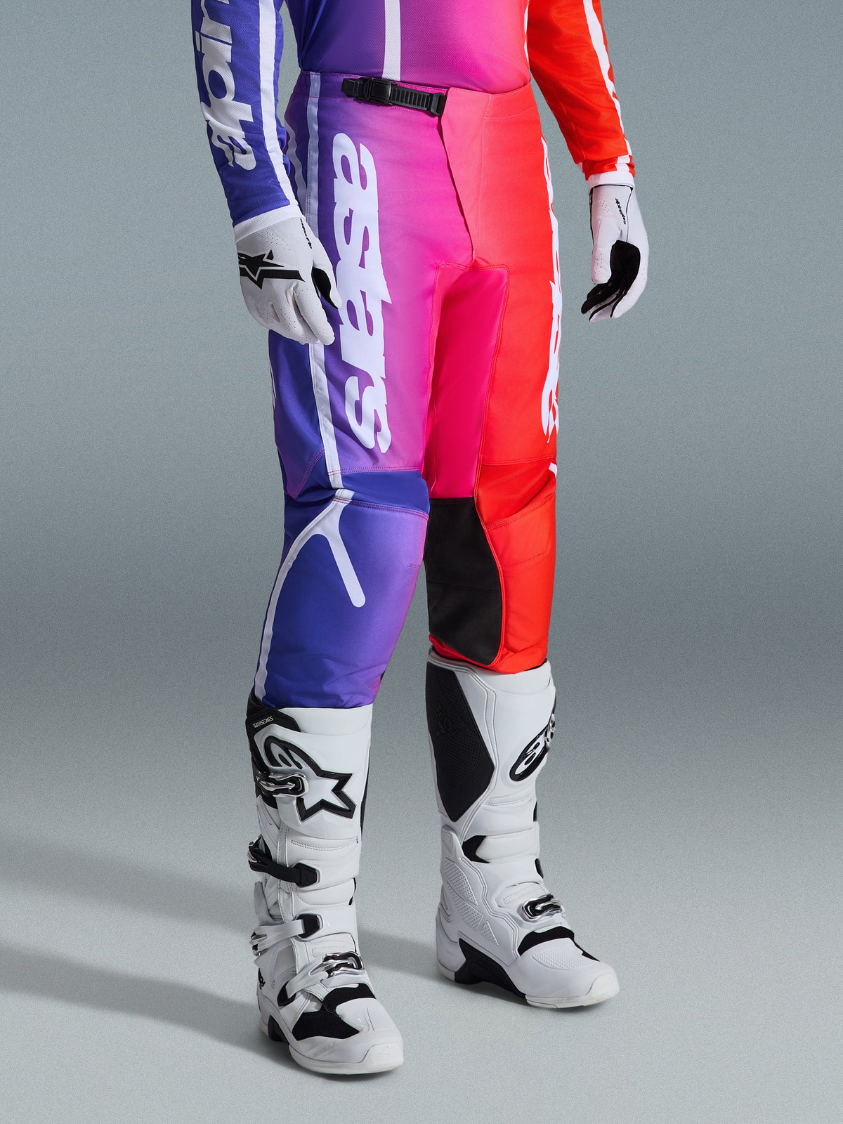 Pantalon Alpinestars Fluid Apex 2026, pantalon de motocross, dégradé multicolore violet, rose et rouge, porté avec des bottes et des gants de motocross blancs et noirs, avec marquage vertical blanc et accents noirs aux genoux, équipement de performance tout-terrain