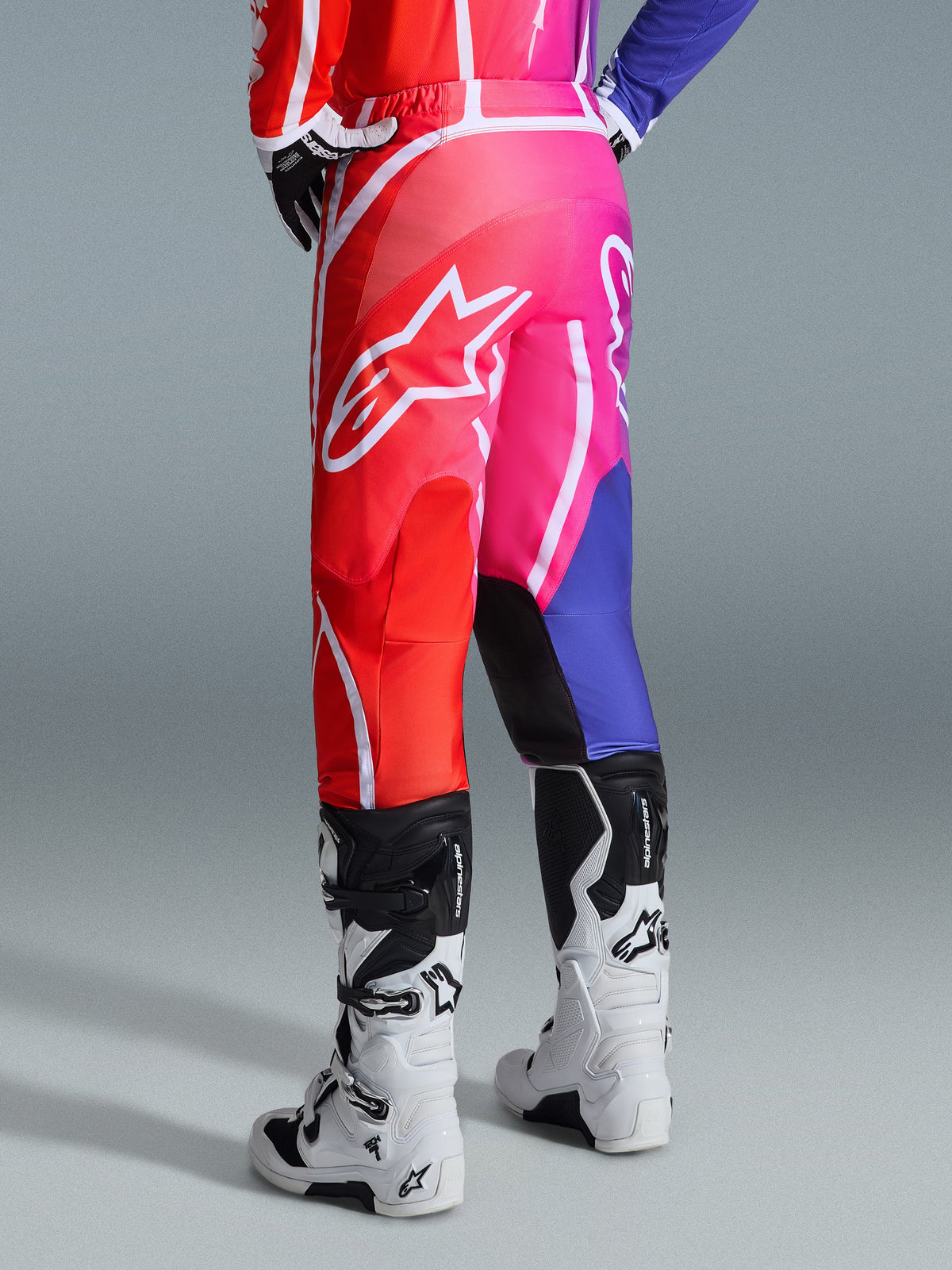 Pantalon Alpinestars Fluid Apex 2026, pantalon de motocross, multicolore rouge, rose et violet, vue arrière d'un pilote portant le pantalon avec des bottes de motocross blanches et noires, arborant un grand logo Alpinestars blanc, design dégradé éclatant