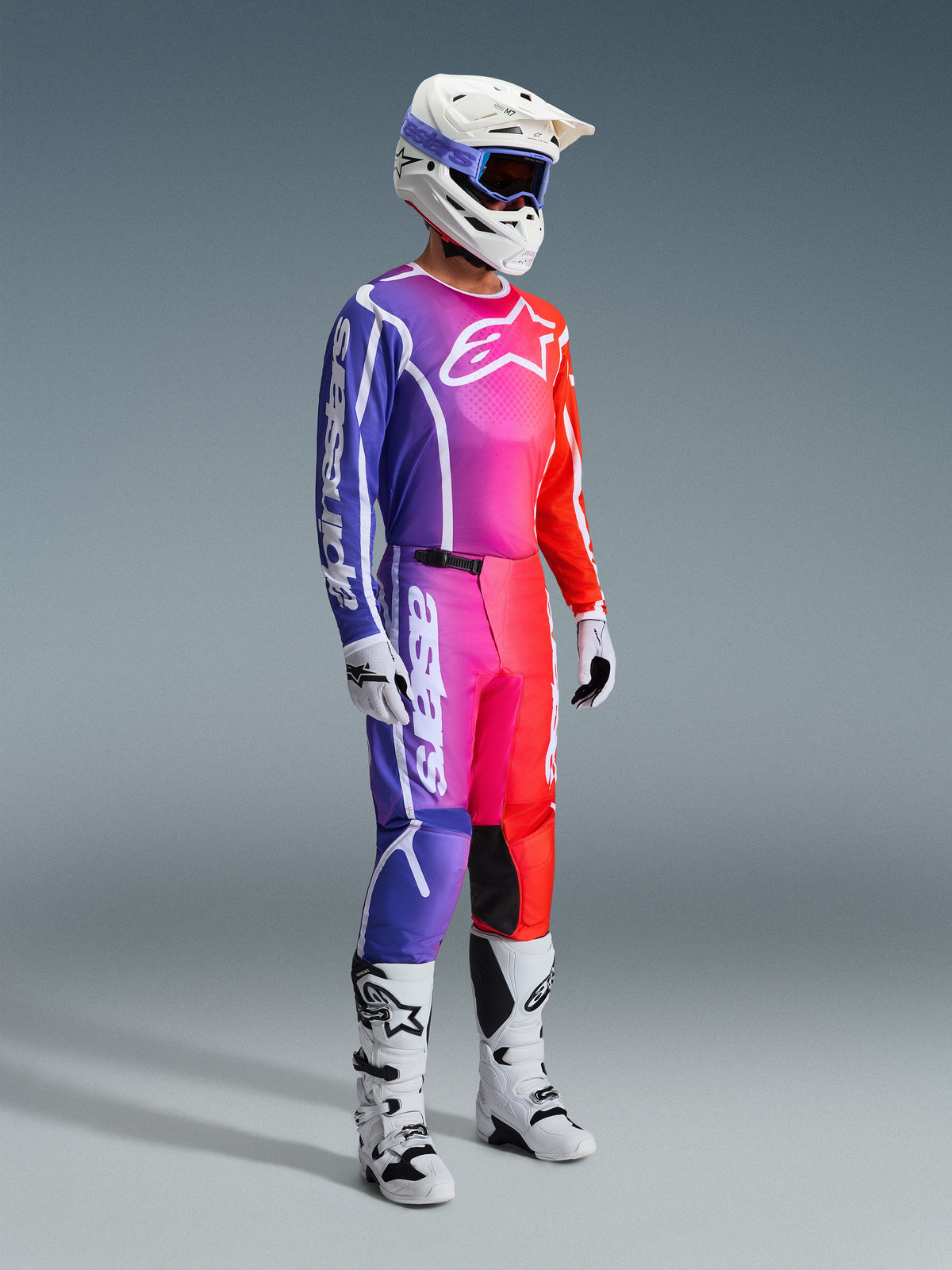 Alpinestars 2026 Fluid Apex Pants, pantalon de motocross, dégradé multicolore de violet, rose et orange, porté avec un maillot assorti, un casque blanc et des bottes blanches, avec un marquage blanc audacieux et un design ergonomique pour la course tout-terrain