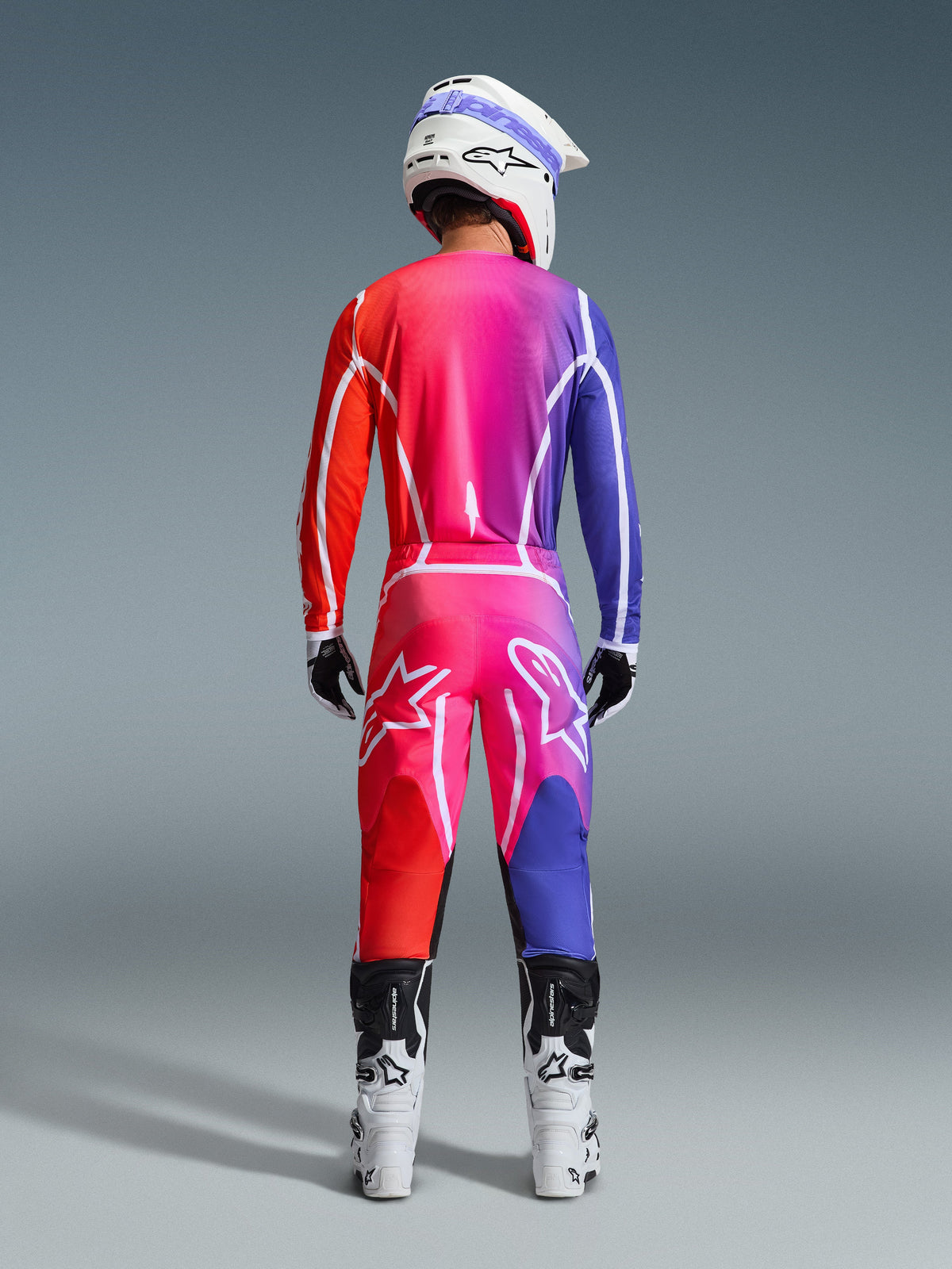 Vue arrière d'un pilote de motocross portant le pantalon Alpinestars Fluid Apex 2026 et le maillot assorti, présentant un dégradé éclatant de rouge, rose et violet, associé à un casque blanc et des bottes blanches, ensemble complet d'équipement coordonné pour la course tout-terrain.