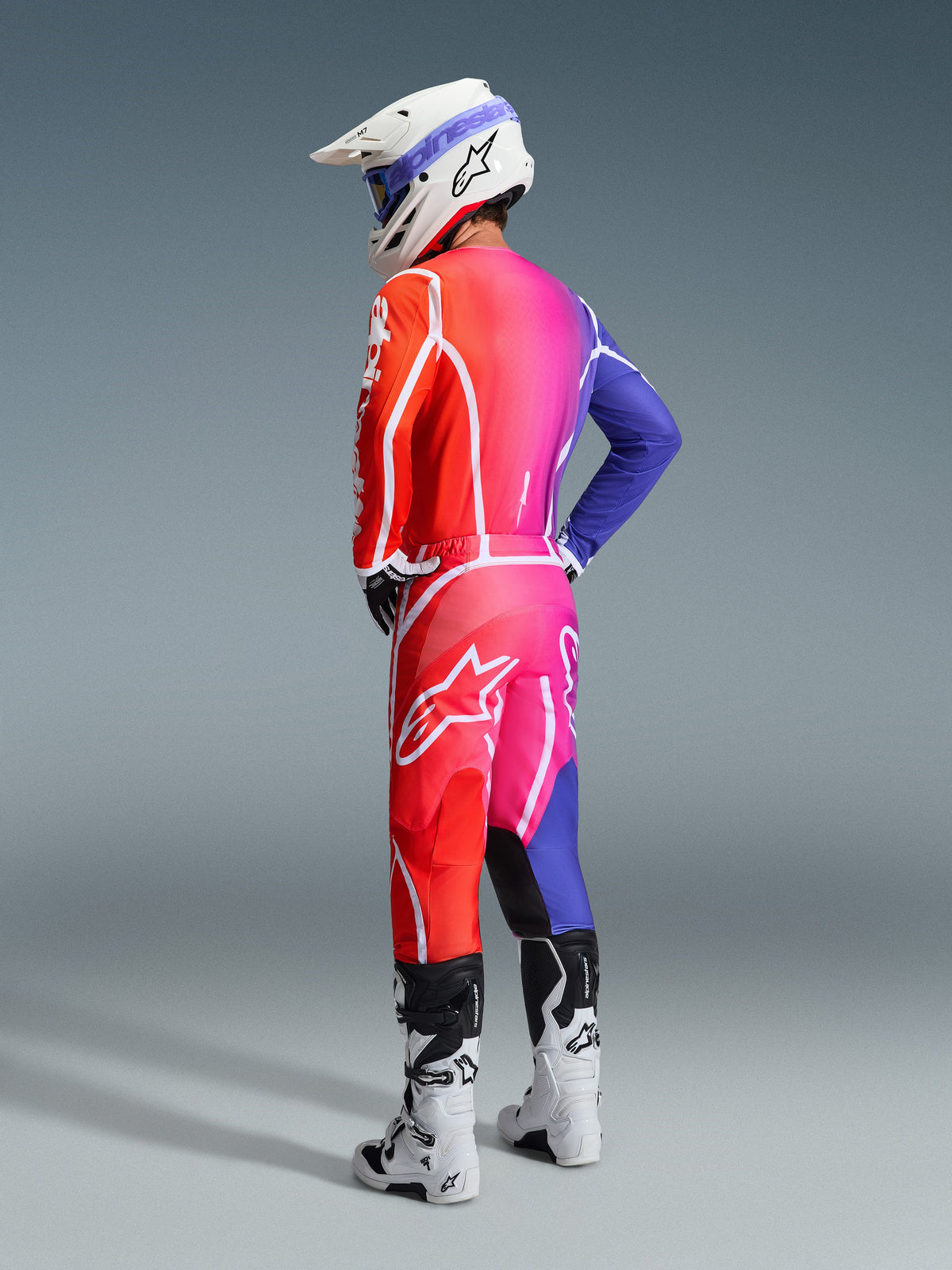 Pilote de motocross portant la tenue Alpinestars Fluid Apex 2026, maillot et pantalon multicolores avec un dégradé rouge, rose et violet et des logos blancs, casque blanc et bottes blanches, tenue coordonnée pour la course tout-terrain