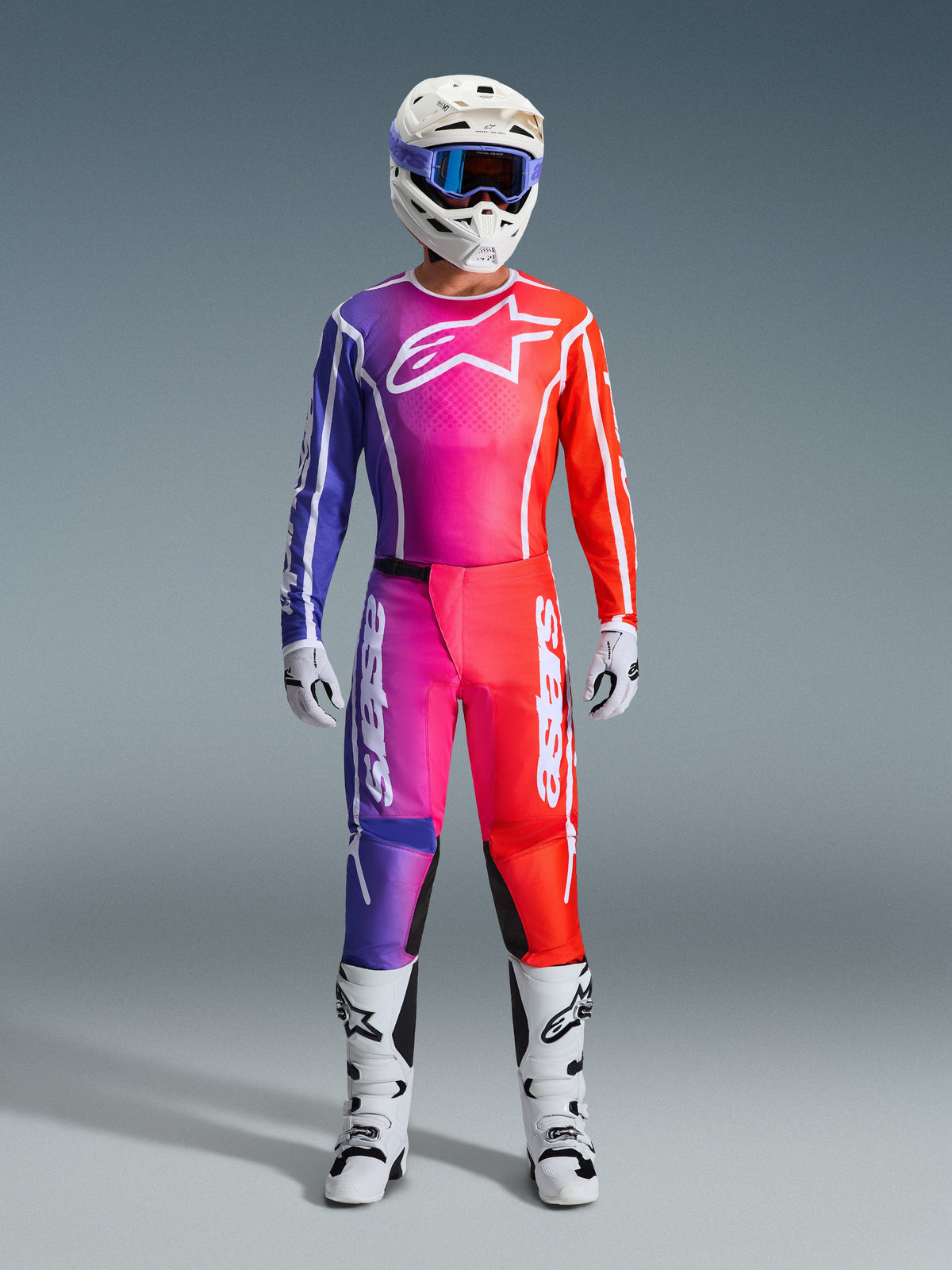 Pilote de motocross portant un pantalon Fluid Apex 2026 et un maillot assorti, dégradé violet et orange, casque blanc avec lunettes, bottes et gants blancs, tenue de course Alpinestars coordonnée pour la performance tout-terrain