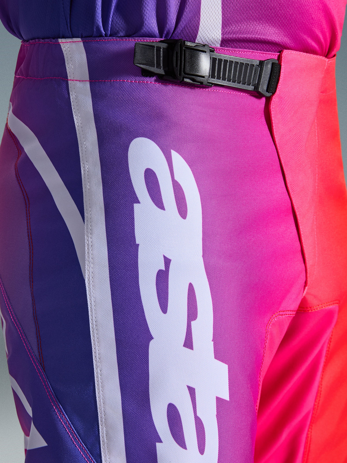 Alpinestars 2026 Fluid Apex Pants, pantalon de motocross, multicolore violet, rose et rouge, gros plan sur la zone de la taille et des hanches avec système de fermeture à cliquet noir et grand logo Alpinestars vertical blanc, design en dégradé éclatant et texture en poly-tissu durable