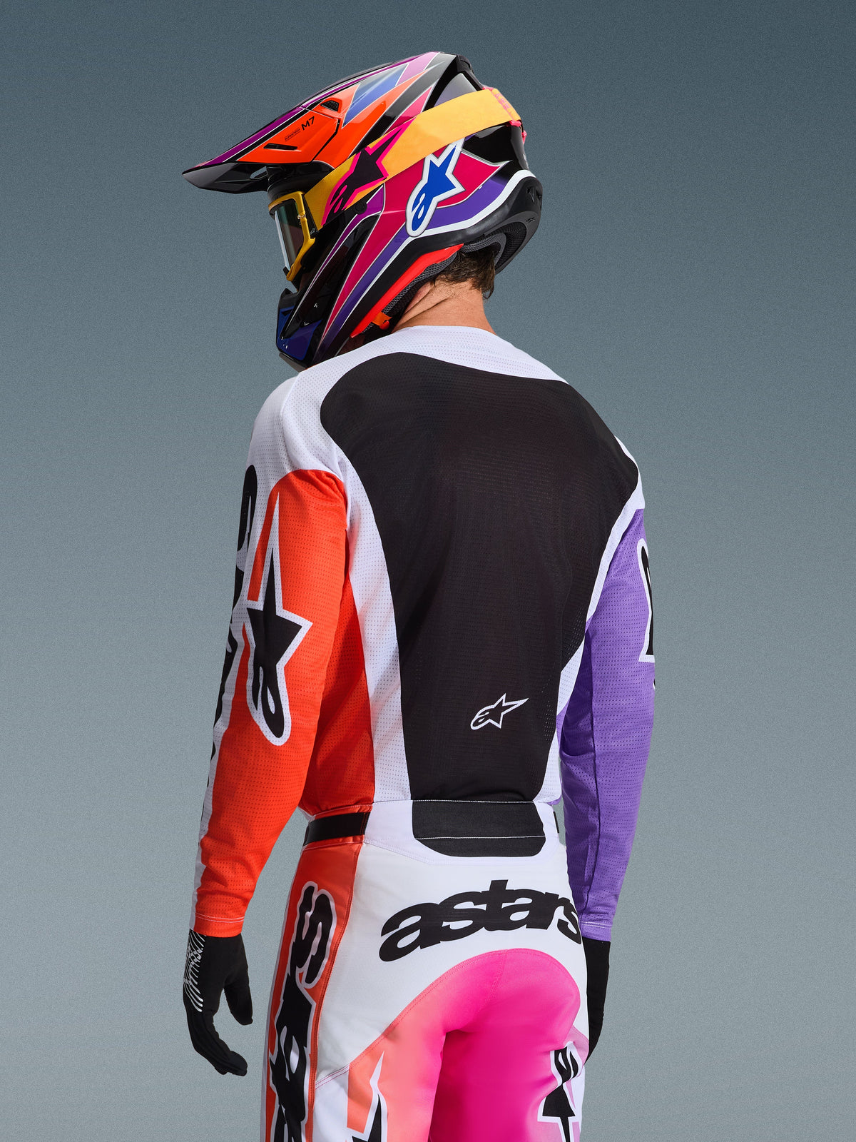 Pilote de motocross de dos portant le maillot Alpinestars 2026 Racer Air Portl Multicolore, avec une manche droite orange, une manche gauche violette et un large panneau arrière en mesh noir pour la ventilation, associé à un pantalon assorti et un casque multicolore éclatant, équipement professionnel tout-terrain.