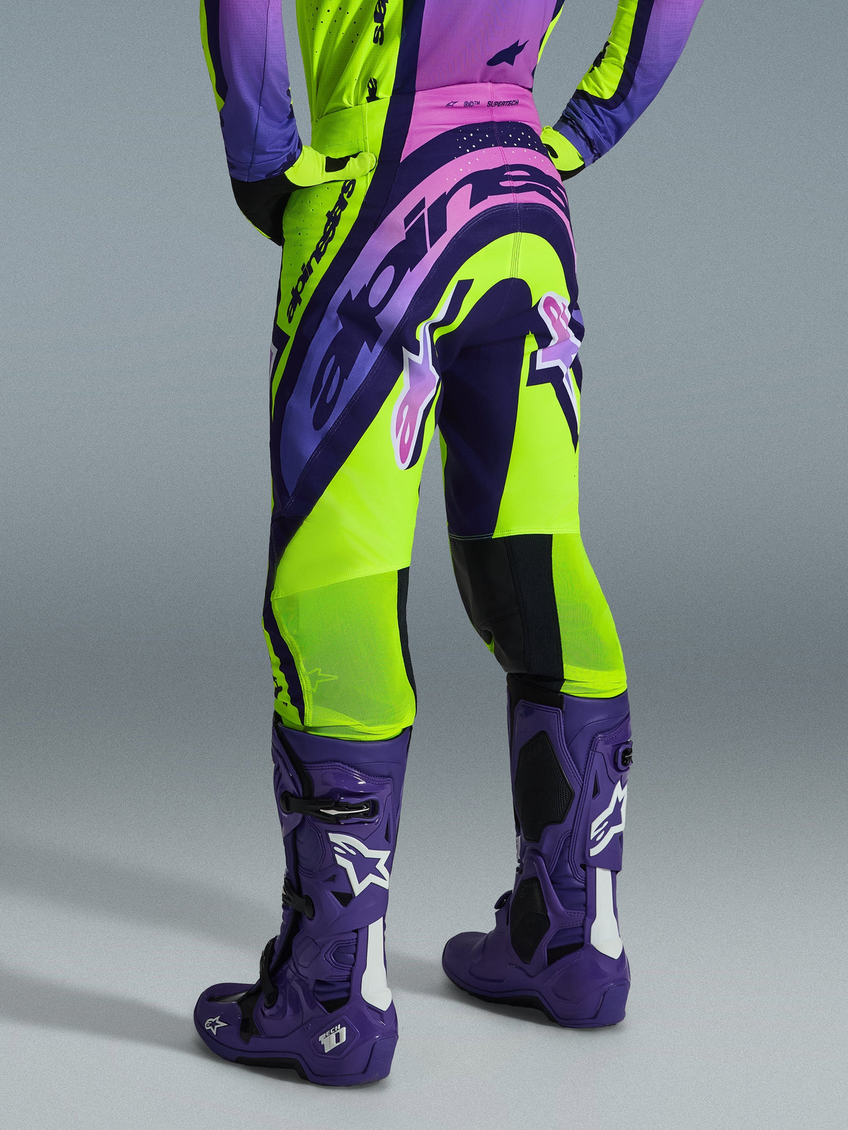 Alpinestars Supertech Vista 2026, pantalon de motocross, jaune, violet et noir, vue arrière porté avec un maillot assorti et des bottes de motocross violettes, doté d'un marquage audacieux et de zones en mesh respirant sur les mollets