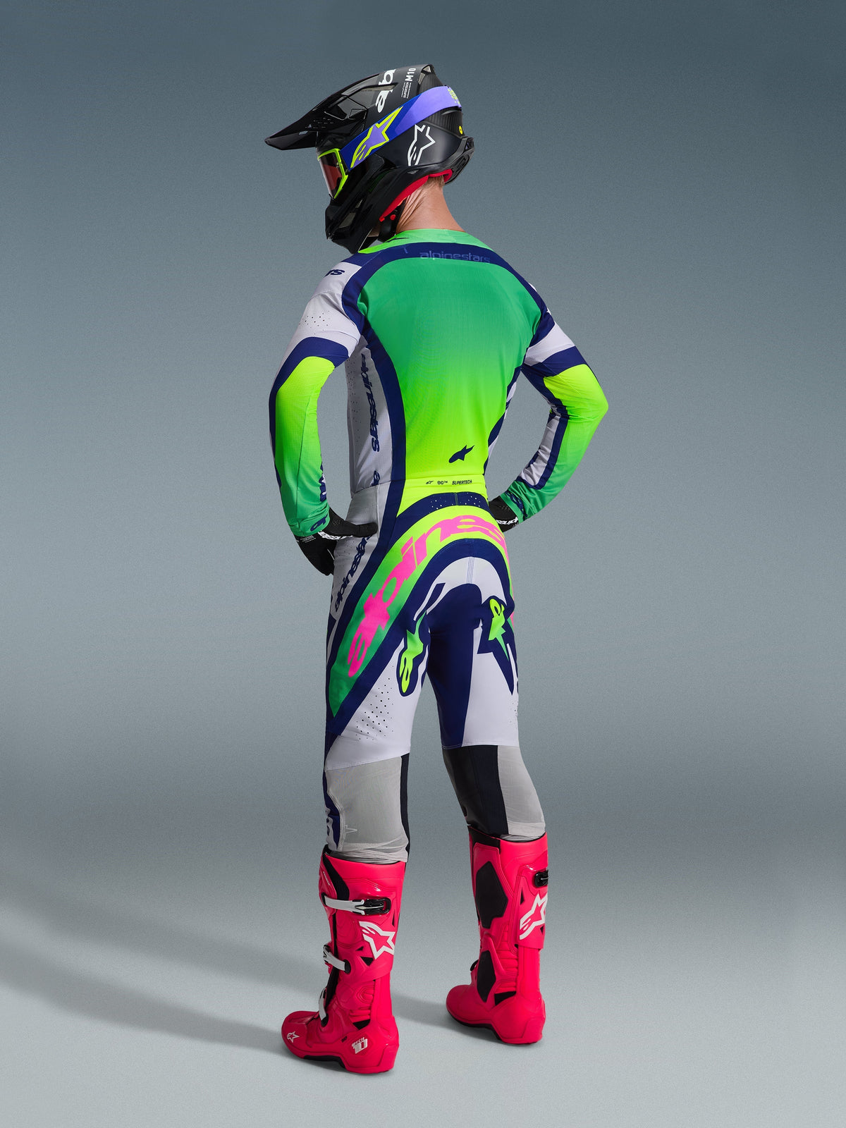 Motocross rider from the rear wearing Alpinestars 2026 Supertech Vista Pants, Light Gray, Blue, and Neon Yellow design with a bold pink logo on the seat, paired with a matching jersey, black helmet, and bright pink boots for a complete off-road racing kit.