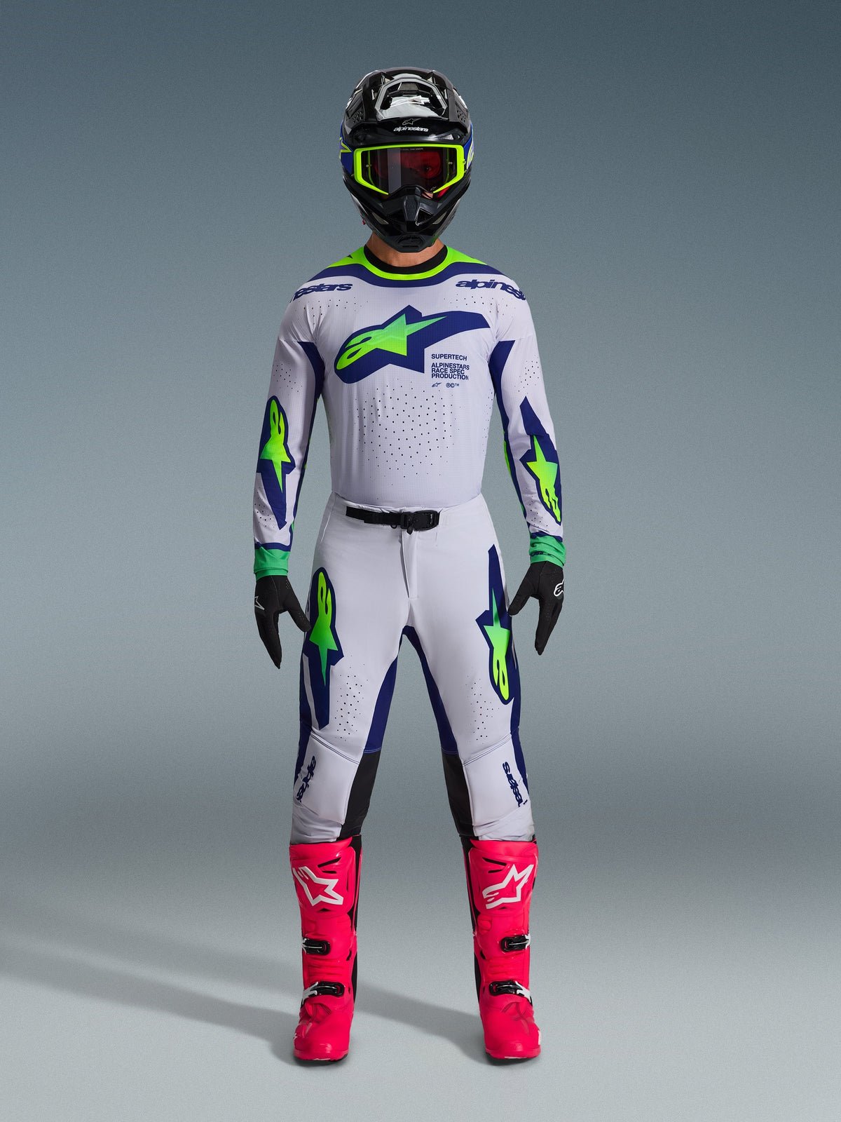 Motocross rider wearing Alpinestars 2026 Supertech Vista Pants and matching jersey, Light Gray with Yellow Fluo and Blue accents, athletic fit, paired with a black helmet, yellow goggles, and vibrant pink boots, professional gear set for off-road racing