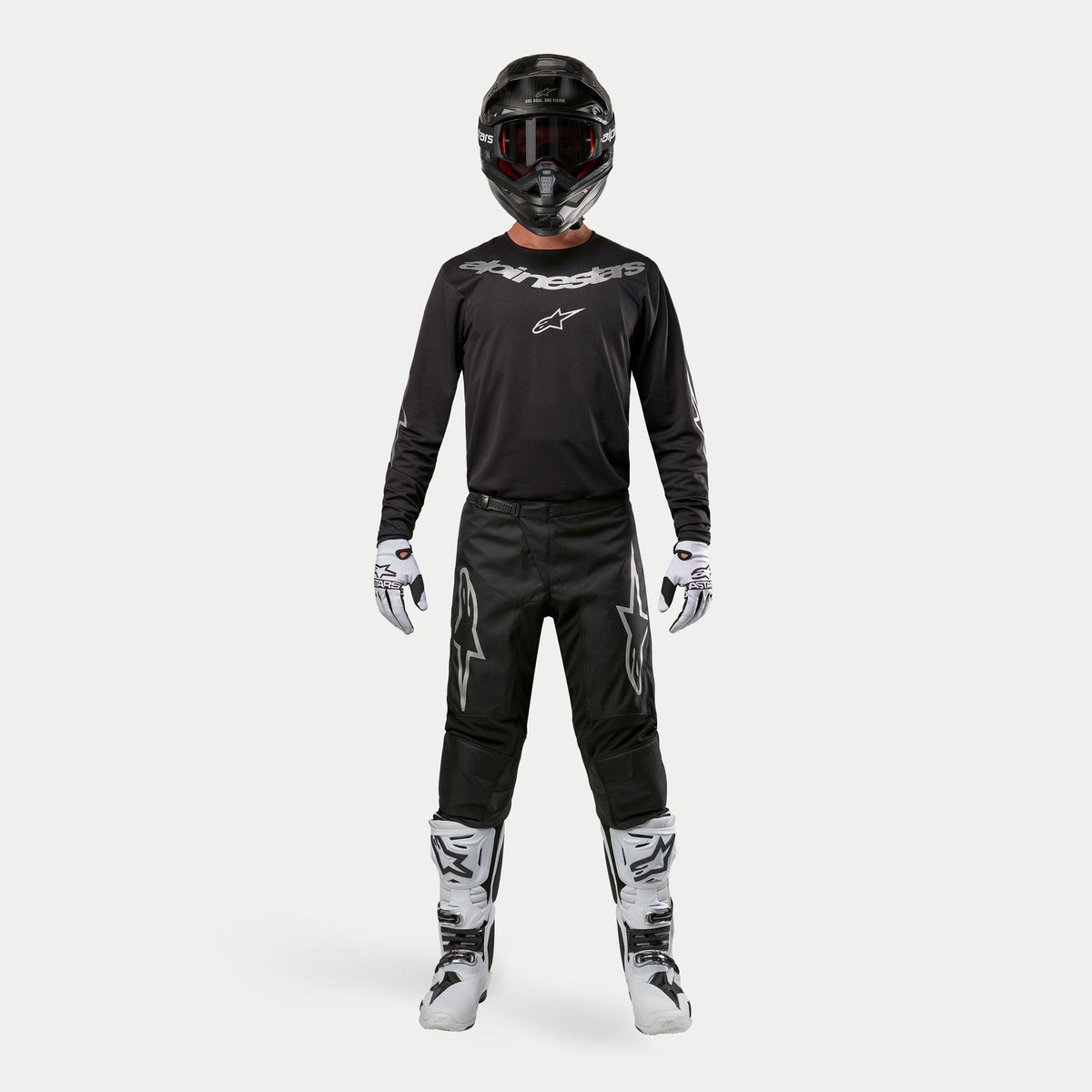 Motocross rider wearing complete racing gear, Alpinestars 2024 Fluid Graphite Pants in Black and Silver, black long-sleeve jersey with silver branding, black helmet with goggles, white and black boots, white and black gloves, coordinated outfit for motocross