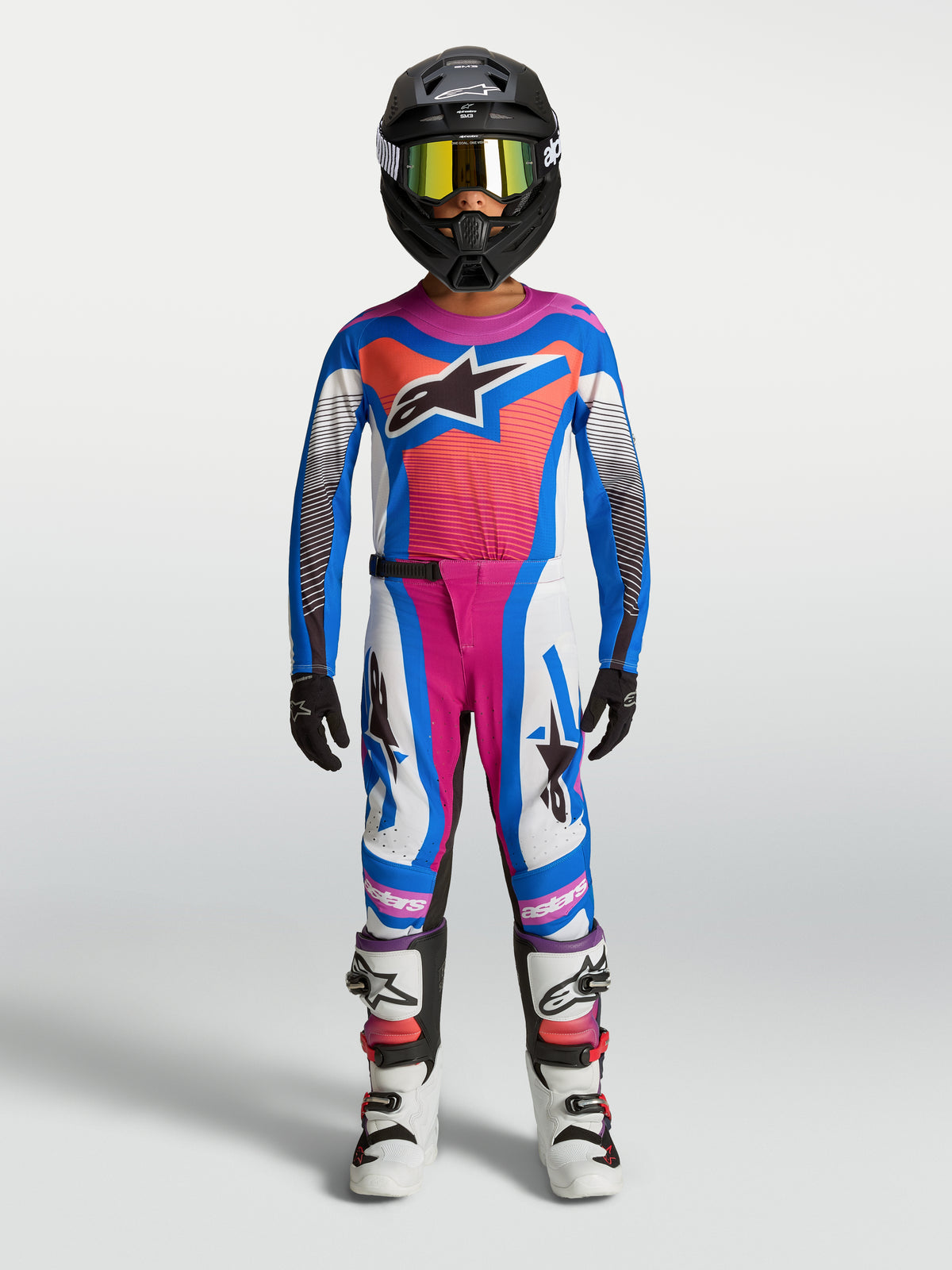 Pantalon Supertech Maven Junior Alpinestars, équipement de motocross pour enfant, blanc, rose et bleu, coupe athlétique pour la course tout-terrain, porté par un jeune pilote avec maillot et casque assortis