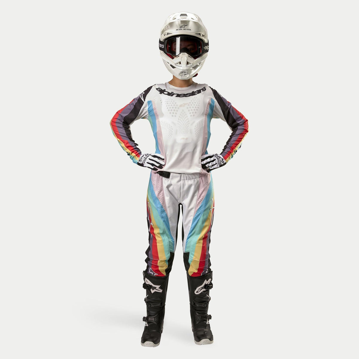 Pilote de motocross portant le pantalon Alpinestars Stella Techstar 2024 pour femme, Noir Multicolore avec base blanche et rayures rouges, jaunes et bleues, maillot à manches longues assorti, casque blanc et bottes noires, tenue de course complète pour la compétition tout-terrain