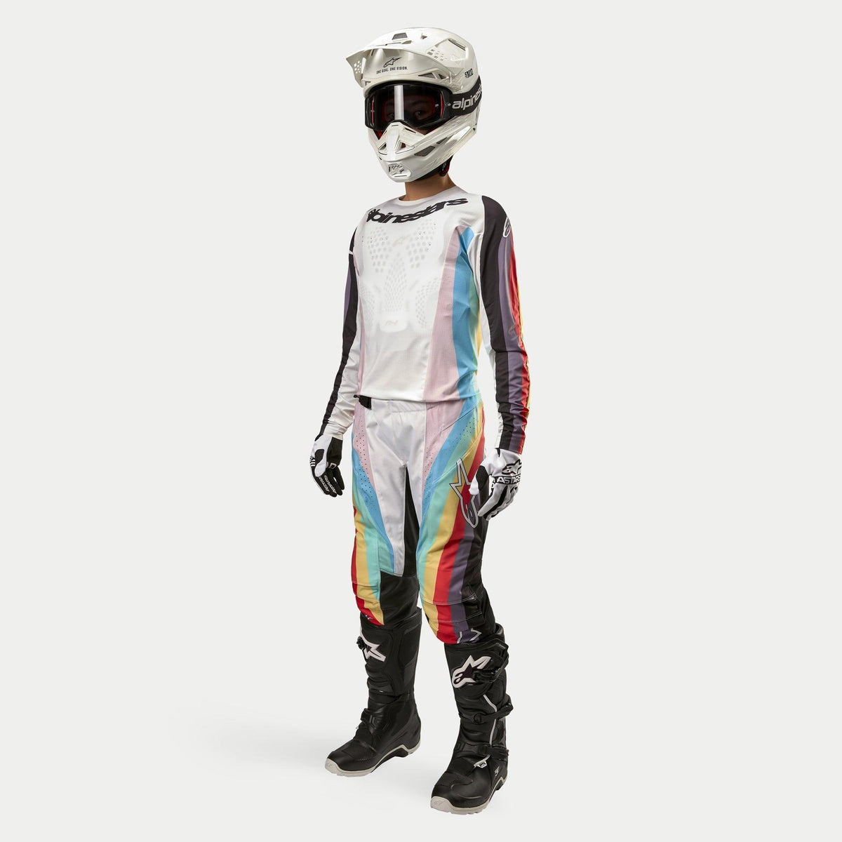 Pilote de motocross portant le pantalon femme Alpinestars Stella Techstar 2024, pantalon de motocross, noir multicolore avec empiècements blancs et rayures diagonales vives, maillot assorti, casque blanc et bottes noires, tenue de course tout-terrain coordonnée pour femme