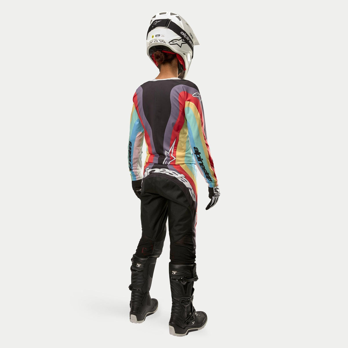 Pilote de motocross vue de dos portant le pantalon Alpinestars Stella Techstar 2024 pour femme en Noir Multicolore, avec un grand marquage blanc sur l'assise, assorti à un maillot à rayures colorées, un casque blanc et des bottes Tech 3 noires pour la course tout-terrain.