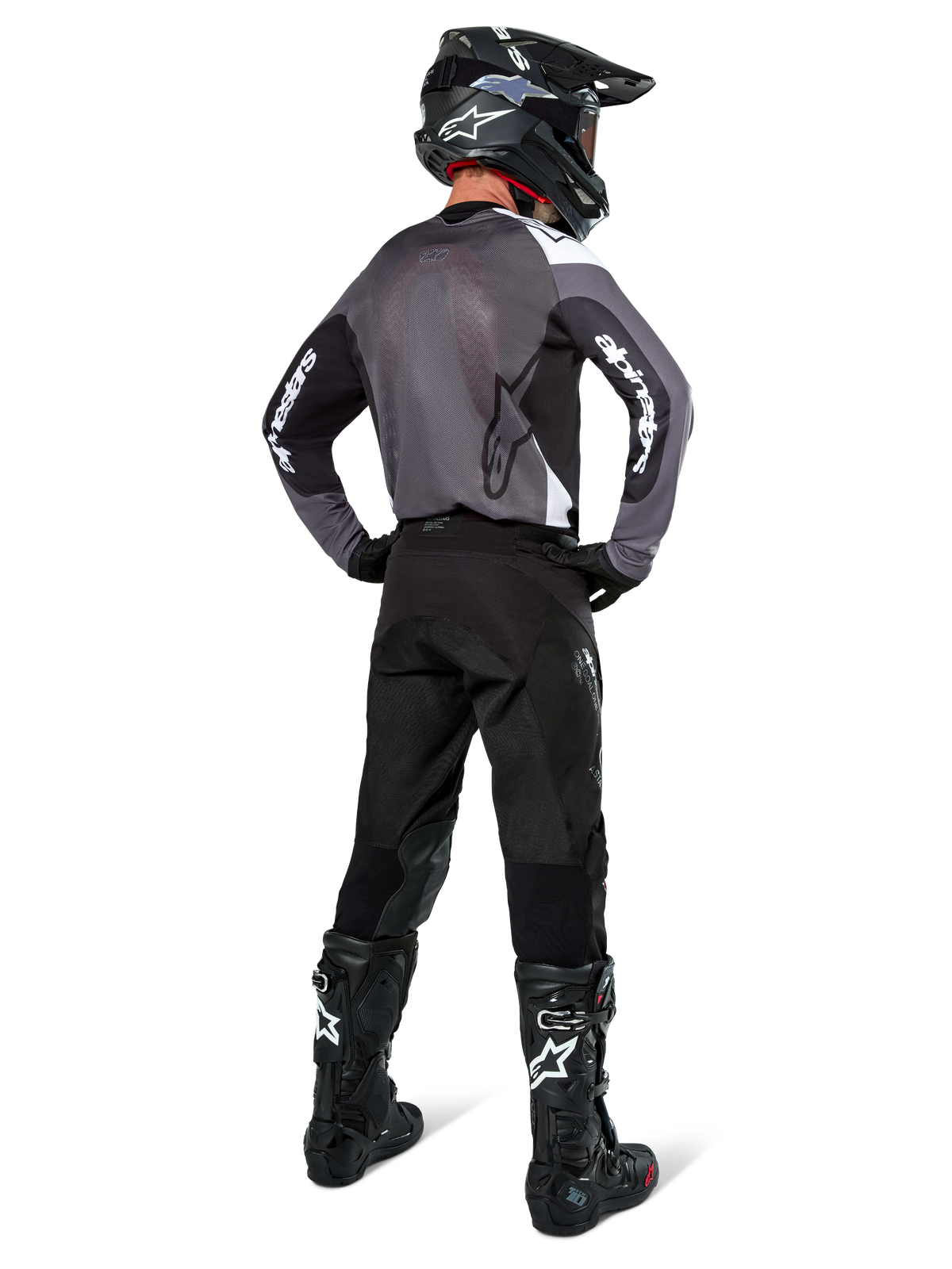 Motocross rider wearing Alpinestars Pro-Dura Jersey in Black Magnet White, rear view showing grey mesh back panel and black sleeves with white Alpinestars branding, coordinated with black pants, black helmet, and black and white boots, professional enduro racing gear