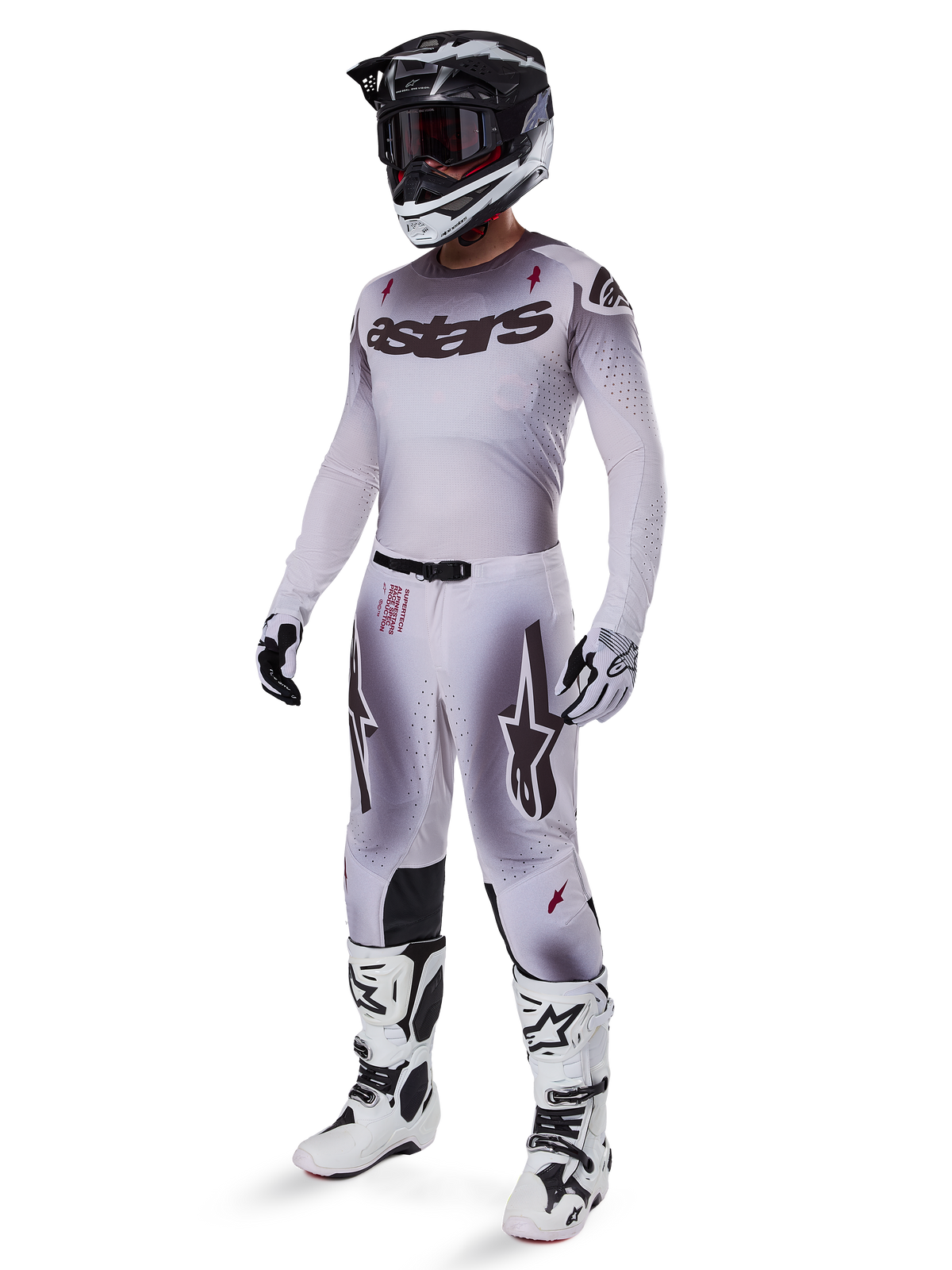 Pilote de motocross portant une tenue de course complète, maillot avec dégradé gris et blanc et logo 'astars' noir, pantalon assorti avec accents rouges et logos étoiles, casque noir et blanc avec lunettes sombres, bottes Alpinestars Tech 10 blanches, tenue de course tout-terrain professionnelle