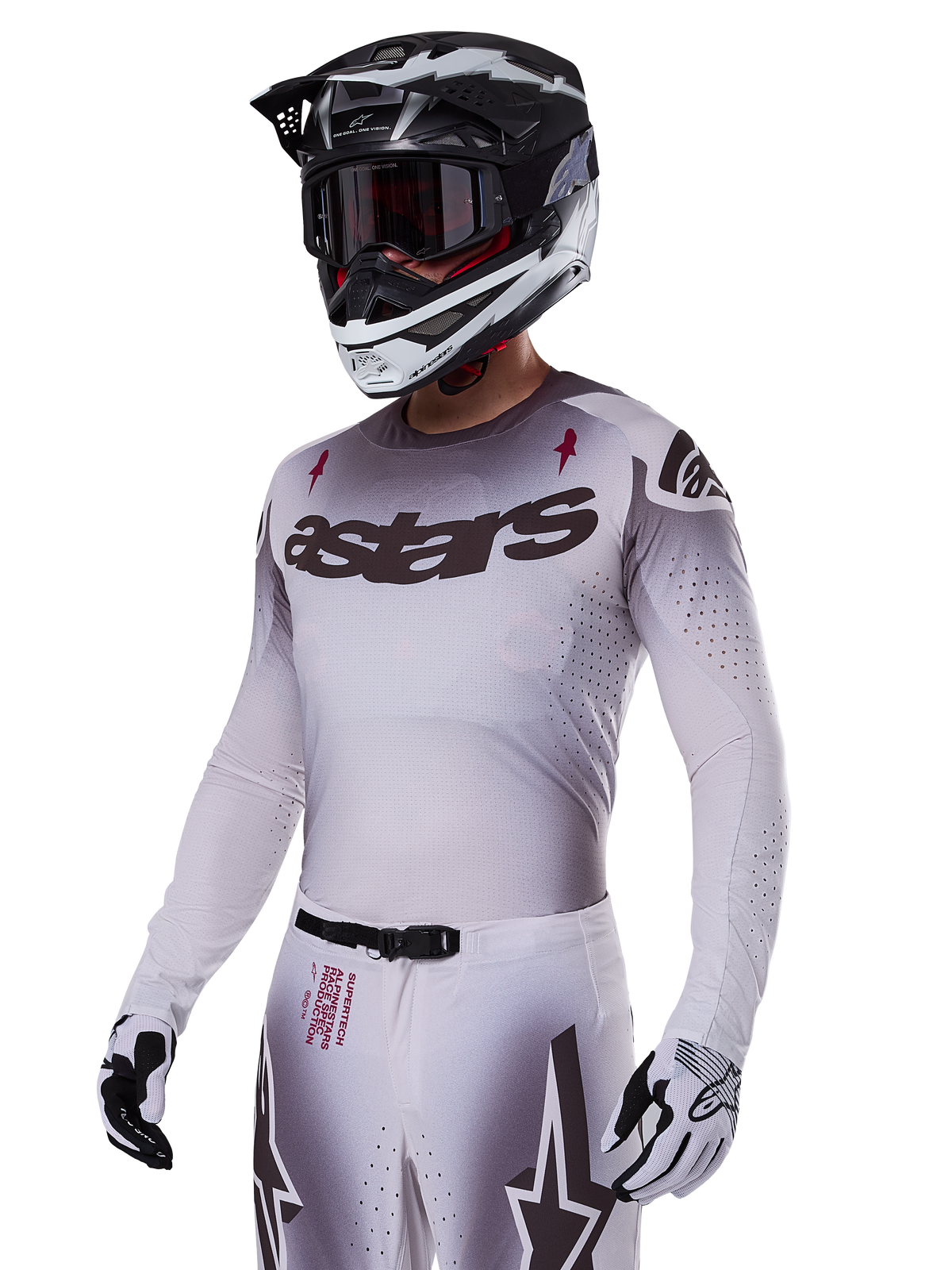 Motocross rider wearing the Alpinestars 2025 Supertech Maker Gear Set in White, featuring a grey gradient jersey with the black astars logo and laser-cut perforations, matching pants with magnetic buckle and star graphics, worn with a black and white helmet and gloves for a complete off-road racing look.