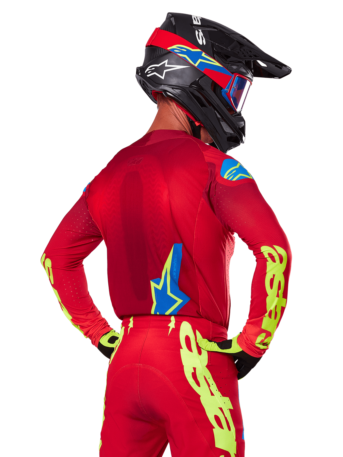 Vue arrière d'un pilote de motocross portant l'ensemble Alpinestars 2025 Supertech Maker rouge, comprenant un maillot rouge avec marquage jaune fluo sur les manches, un pantalon rouge assorti, un casque noir avec des accents bleus et rouges, et des gants noirs et jaunes coordonnés.