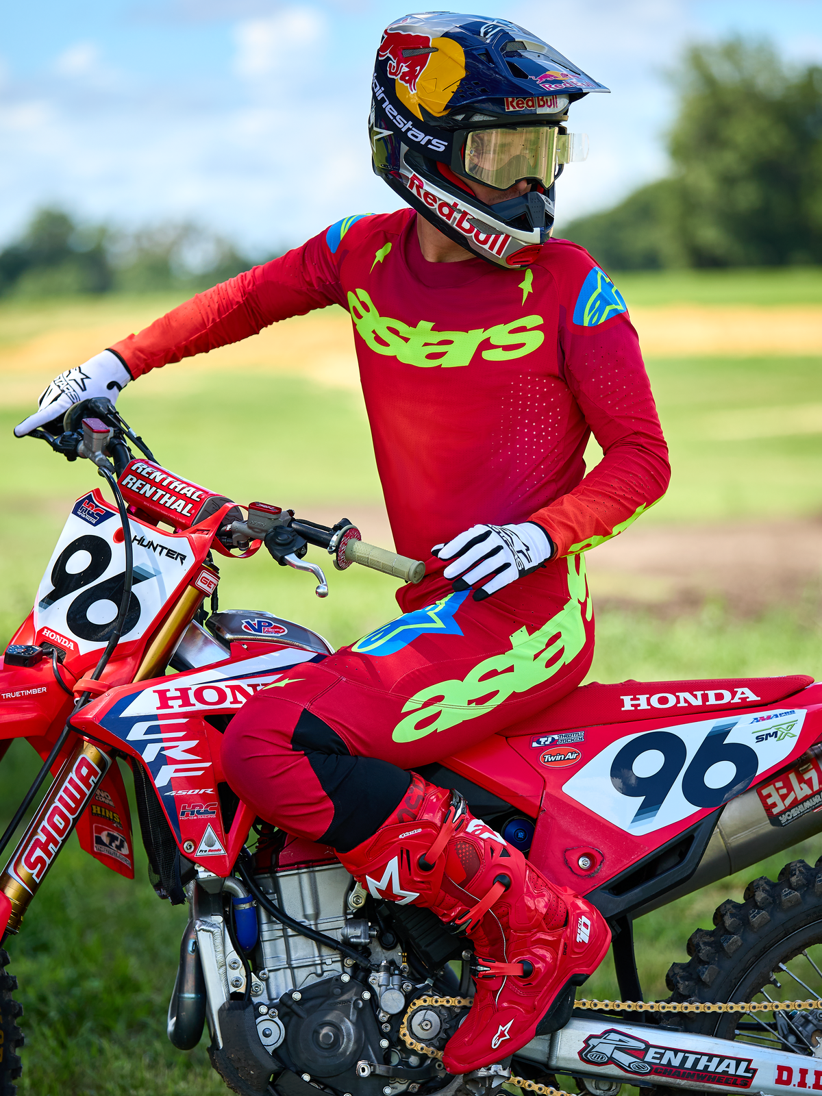 Pilote de motocross sur une moto Honda rouge portant l'ensemble Alpinestars 2025 Supertech Maker rouge, comprenant un maillot rouge avec logo 'astars' vert fluo et accents bleus aux épaules, pantalon rouge assorti, bottes rouges, gants blancs et casque Red Bull, kit de course professionnel pour performances tout-terrain