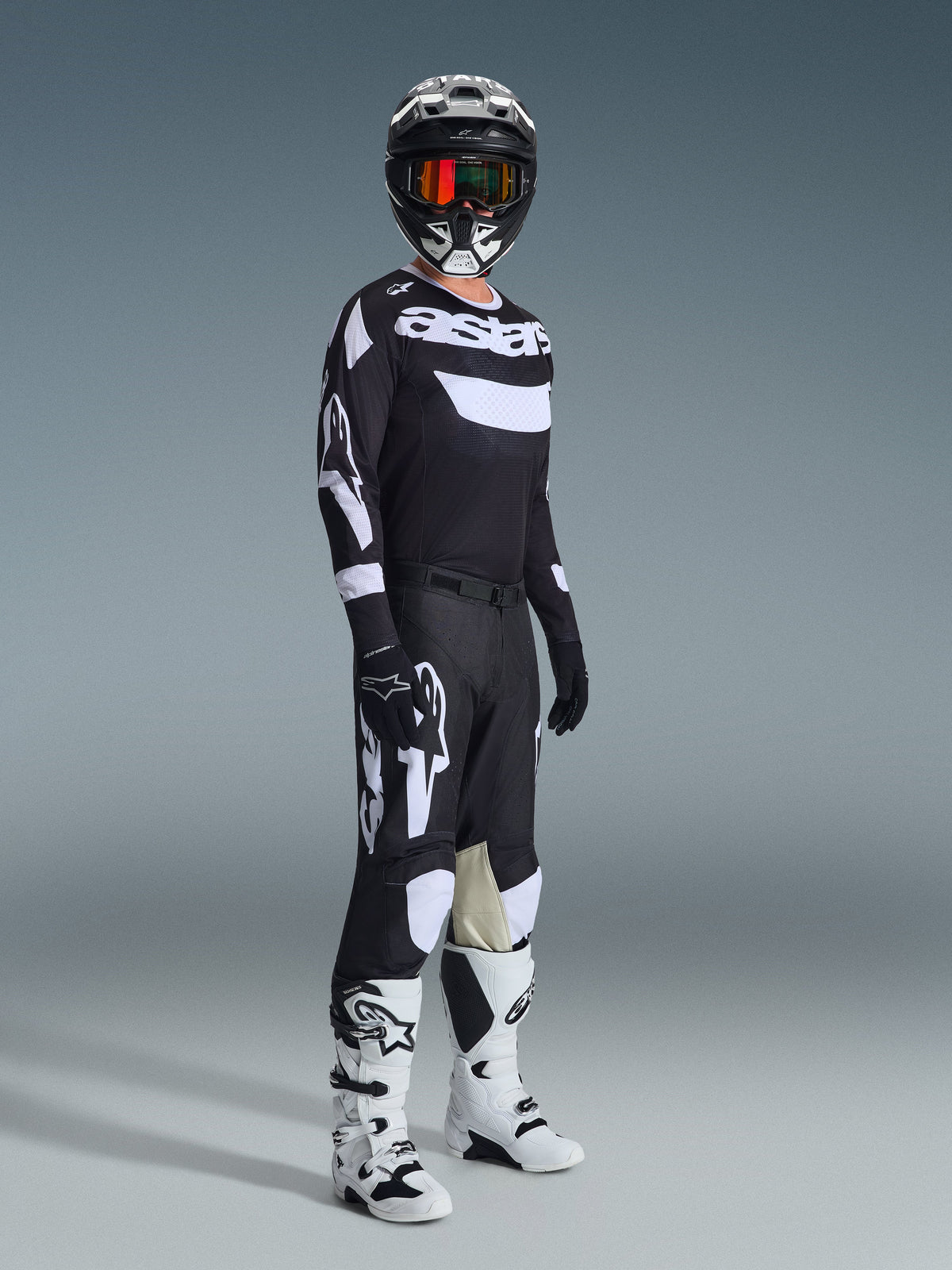 Ensemble d'équipement Alpinestars 2026 Racer Air Riway, pilote de motocross portant une tenue de course complète, maillot noir avec logos 'Astars' blancs et panneaux en mesh, pantalon noir avec grands logos blancs et panneaux aux genoux, casque noir avec lunettes rouges, bottes blanches et noires, tenue de course tout-terrain professionnelle.