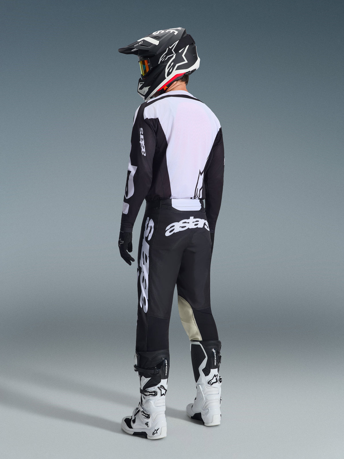 Pilote de motocross vu de dos portant l'ensemble Alpinestars 2026 Racer Air Riway noir et blanc, avec un dos de maillot en maille ventilée et de larges logos blancs, assorti d'un casque noir et de bottes blanches.