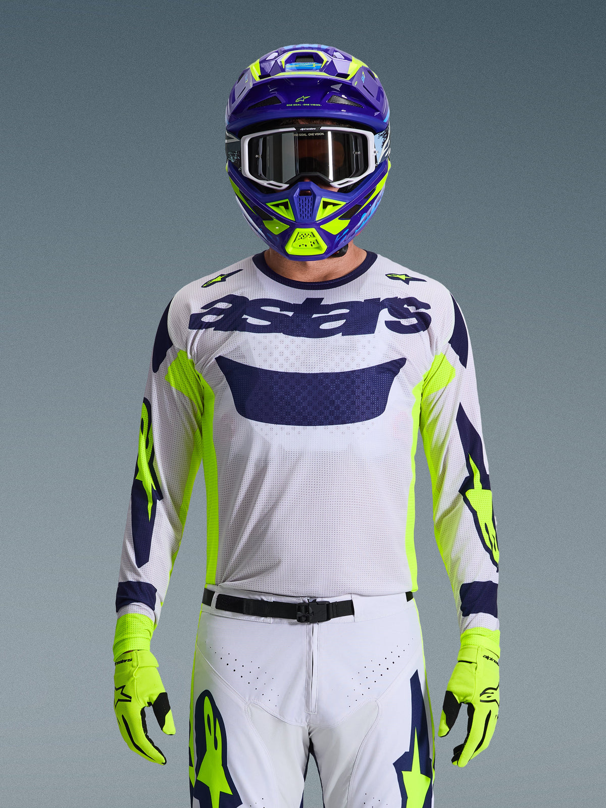 Pilote de motocross portant la tenue Alpinestars 2026 Racer Air Riway, maillot en mesh blanc avec logo 'astars' bleu marine et accents jaune fluo, pantalon blanc assorti avec graphismes bleu marine et jaune fluo, gants jaune fluo, casque violet et jaune fluo avec lunettes blanches, photo de studio d'un équipement de course tout-terrain complet