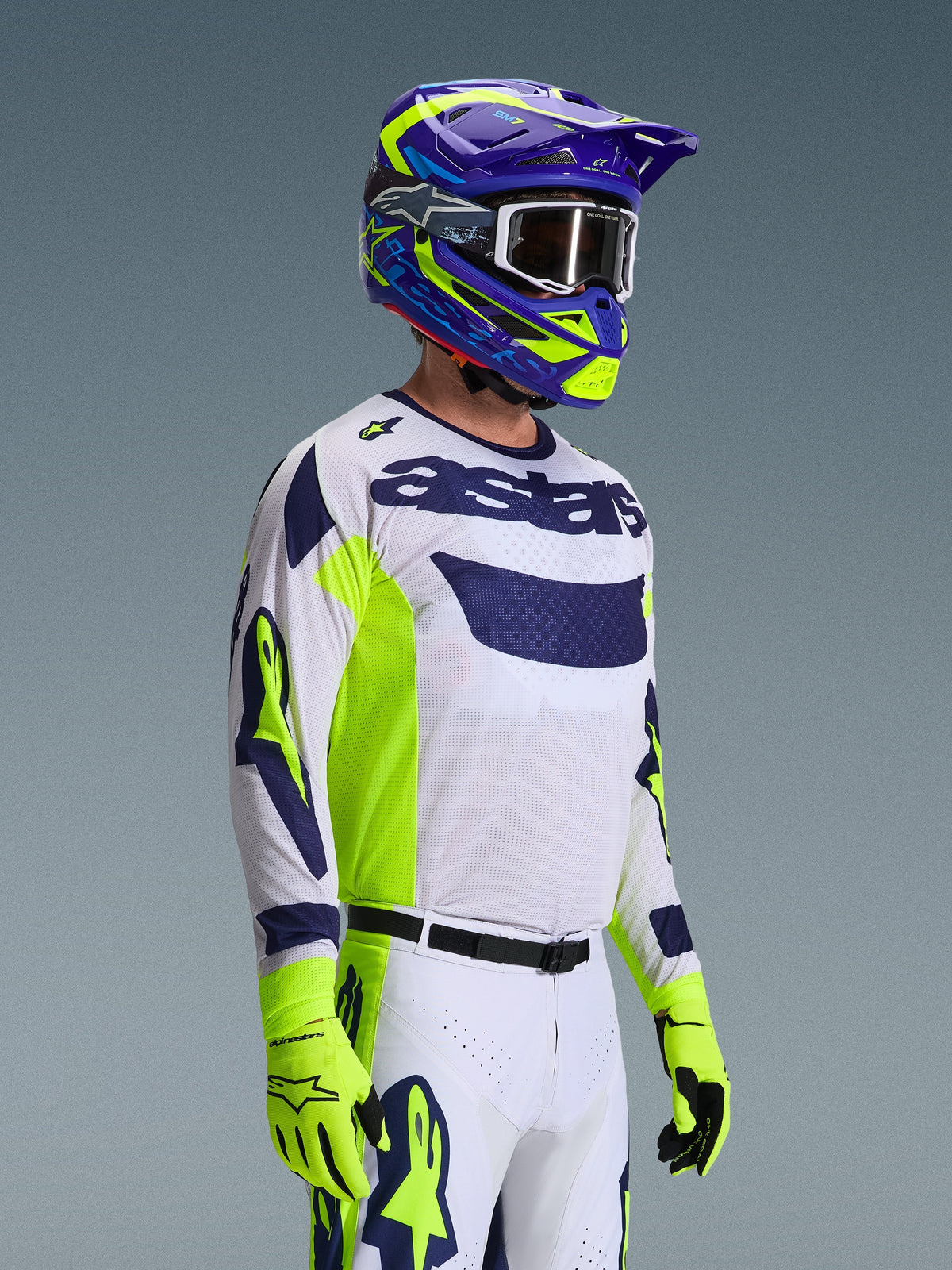Pilote de motocross portant la tenue Alpinestars 2026 Racer Air Riway, maillot blanc avec logo bleu marine et panneaux latéraux jaune fluo, pantalon blanc avec accents assortis bleu marine et jaunes, casque violet et jaune fluo avec lunettes blanches, gants jaune fluo, équipement de course tout-terrain haute performance