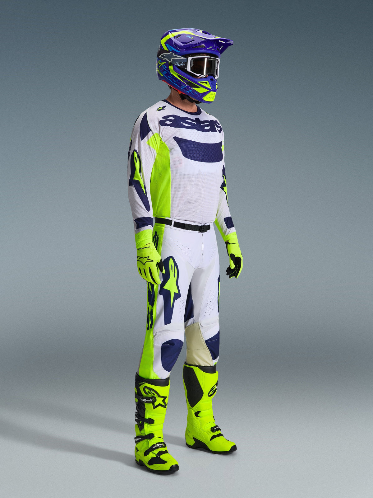 Pilote de motocross Alpinestars portant une tenue de course complète, maillot gris clair avec des accents jaune fluo et bleus, pantalon assorti blanc et jaune, bottes et gants jaune vif, casque violet et jaune avec lunettes foncées, tenue coordonnée pour la course tout-terrain