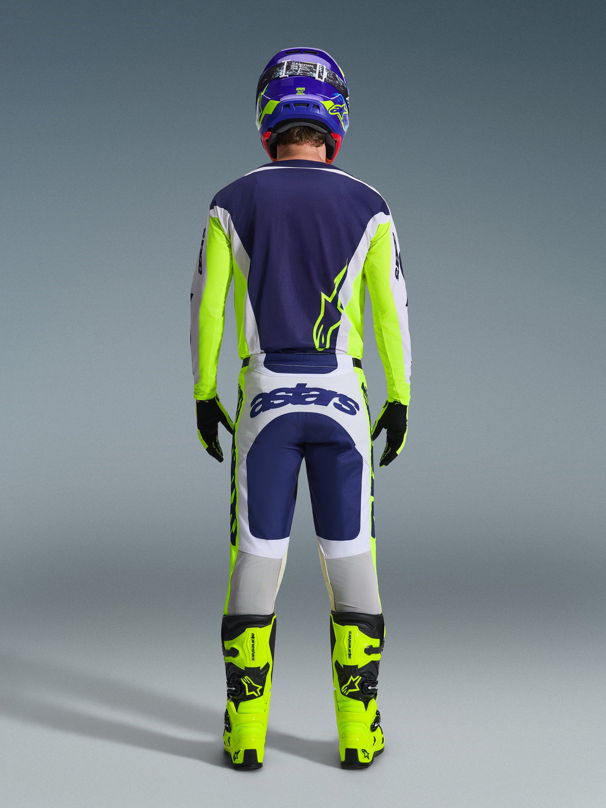 Vue arrière d'un pilote de motocross portant le maillot Alpinestars 2026 Racer Air Riway et le pantalon assorti en gris clair, jaune fluo et bleu, associé à des bottes jaune fluo et un casque coordonné, montrant un équipement de course tout-terrain complet.