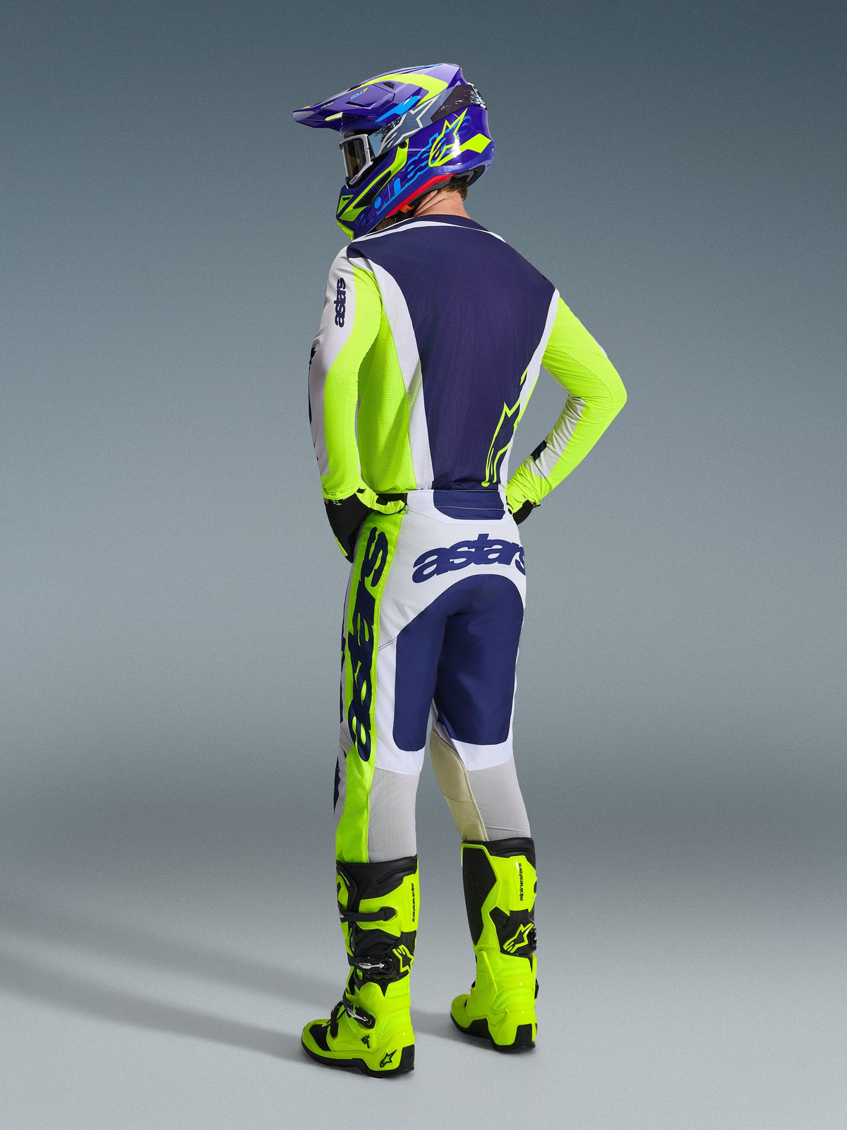 Pilote de motocross vu de dos portant l'ensemble d'équipement Alpinestars Racer Air Riway 2026, maillot et pantalon gris clair, jaune fluo et bleu, avec marquage « astars » sur les manches et l'assise, bottes jaune fluo et casque coloré assorti.