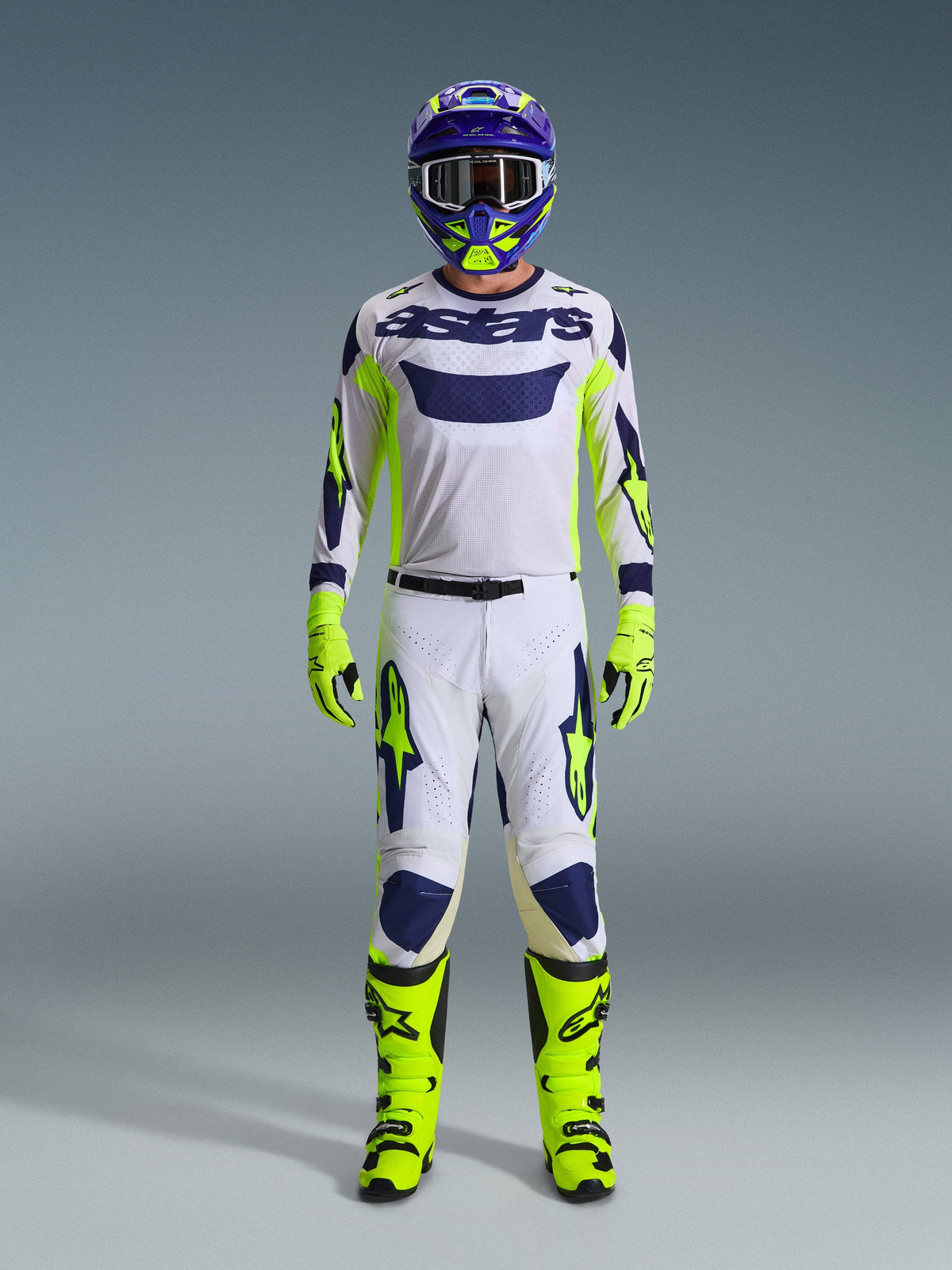 Pilote de motocross en tenue de course complète, maillot Racer Air Riway gris clair avec détails jaune fluo et bleus, pantalon assorti blanc et jaune, bottes et gants jaune fluo, casque violet et jaune, équipement professionnel pour le tout-terrain