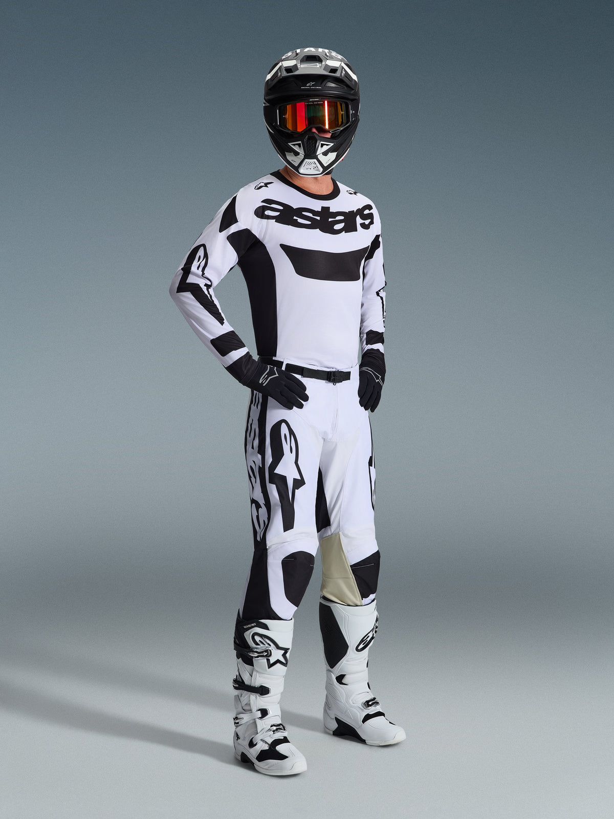 Ensemble d'équipement Alpinestars 2026 Racer Riway, pilote de motocross en tenue de course blanche et noire, maillot blanc avec logo 'astars' noir sur la poitrine, pantalon blanc avec accents noirs, casque noir avec lunettes orange, bottes blanches, équipement coordonné pour le motocross et le tout-terrain