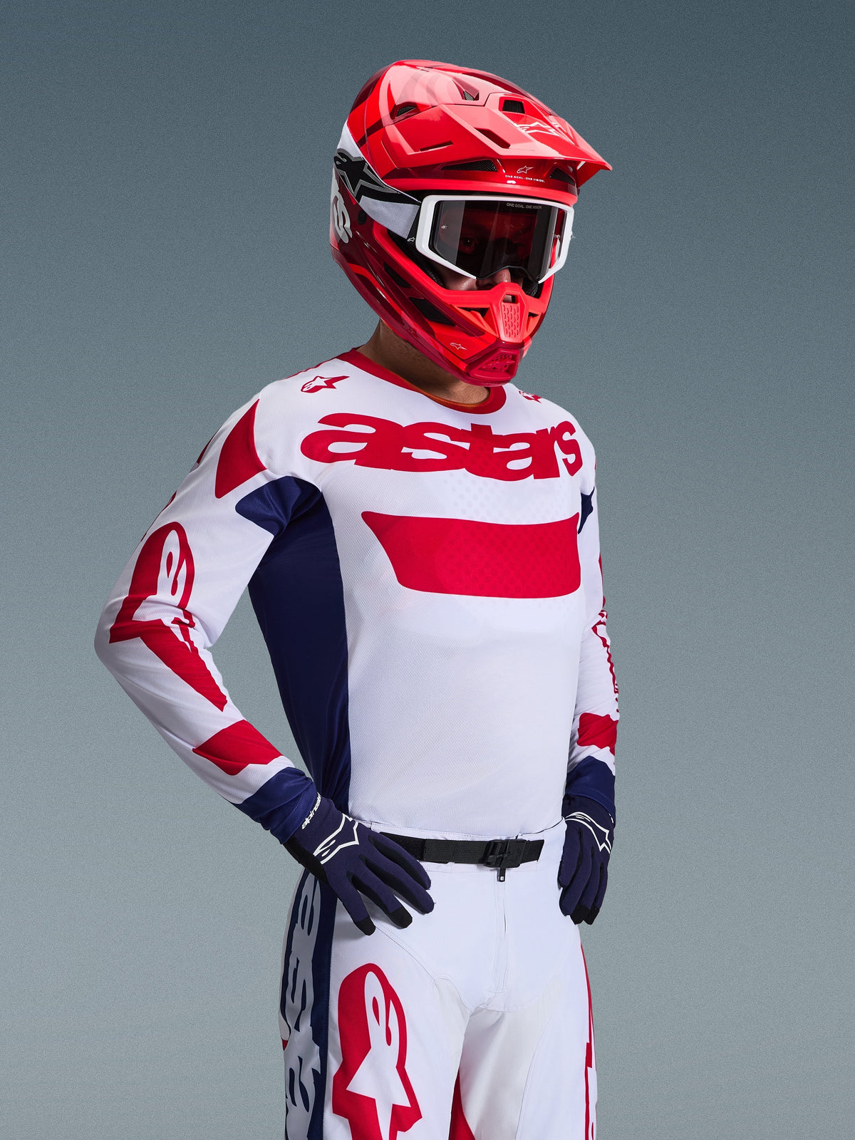 Motocross rider wearing the Alpinestars 2026 Racer Riway Jersey in White Blue Red, matching off-road pants, bright red helmet with white goggles, and navy blue gloves, coordinated high-performance gear set for motocross