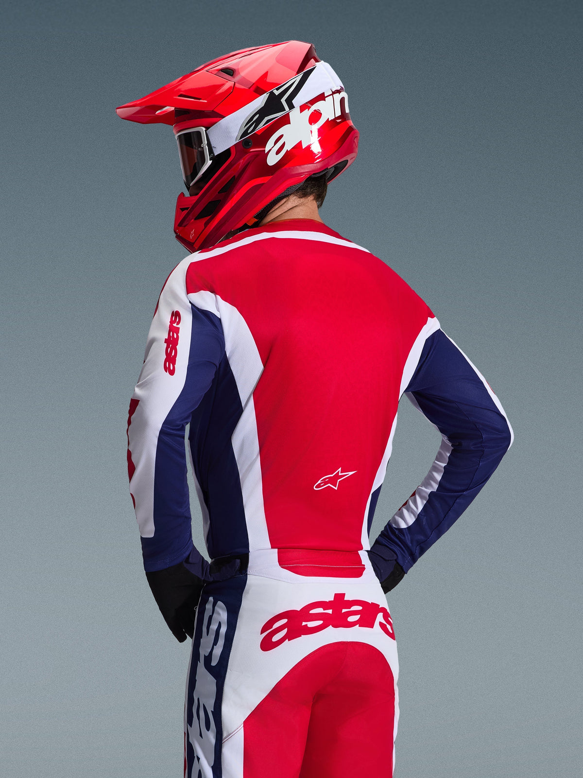 Pilote de motocross de dos portant le maillot Alpinestars Racer Riway 2026, coloris blanc, bleu et rouge, pantalon tout-terrain assorti avec logo astars imposant, casque rouge avec logo Alpinestars blanc, gants noirs, tenue de course de motocross coordonnée