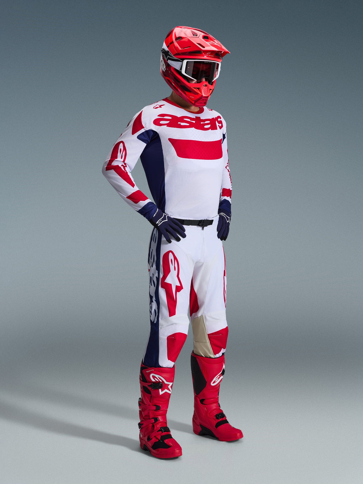 Motocross rider wearing Alpinestars 2026 Racer Riway Gear Set, White jersey with red and blue accents and 'astars' branding, matching white pants with large red logos and navy blue side panels, red helmet, red and black boots, and navy blue gloves, professional racing kit for off-road performance.